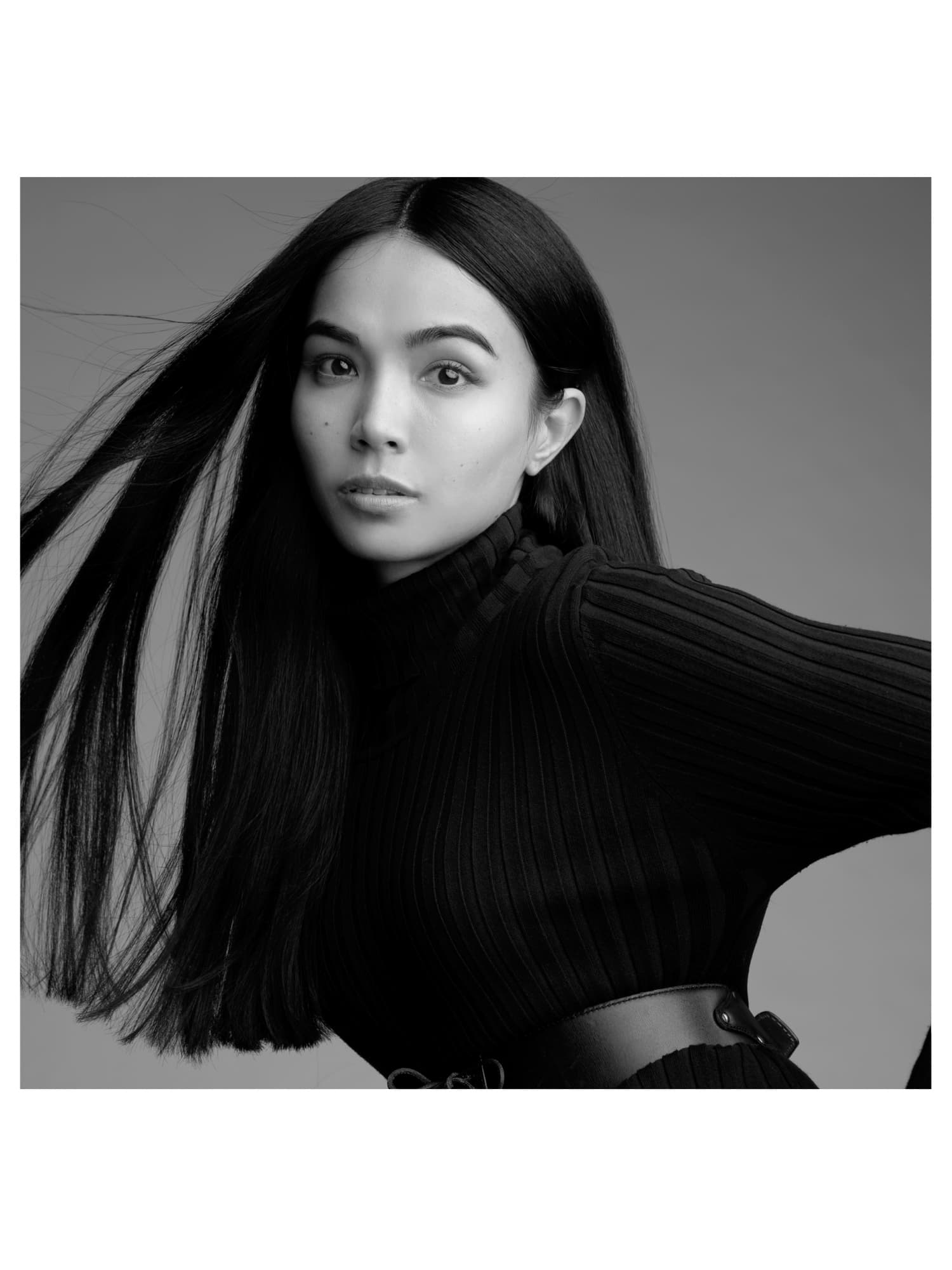 Black-and-white shot of Shirin Hojayeva in a turtleneck and corset belt, tossing her hair with dynamic energy.