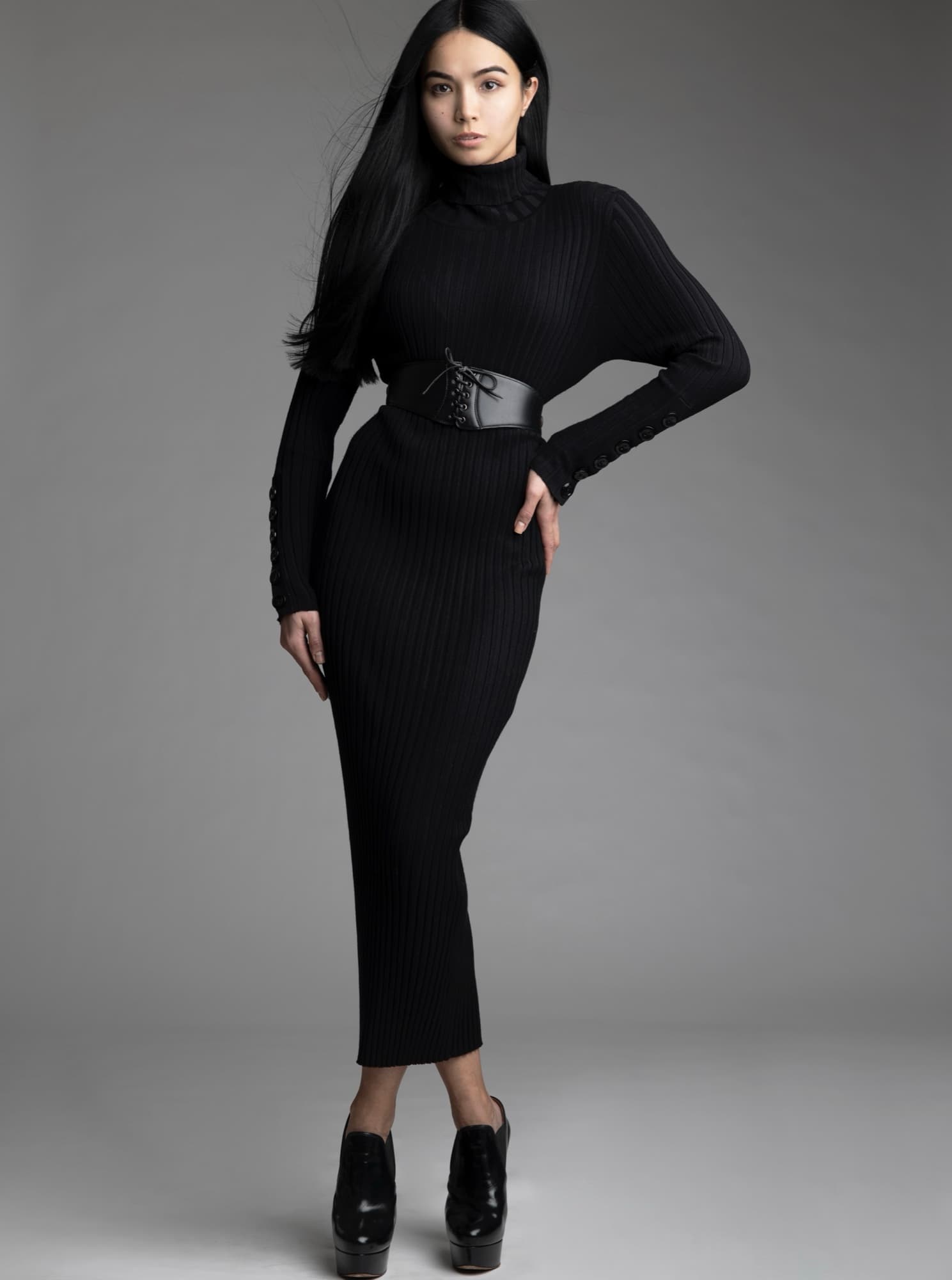 Shirin Hojayeva stands in a black ribbed turtleneck dress with a leather corset belt, striking a poised full-length pose.