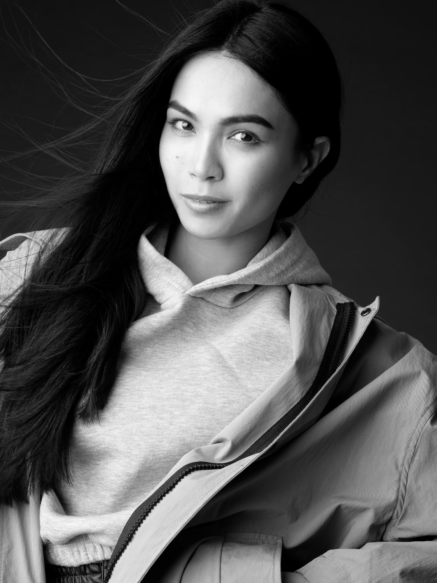 Black-and-white close-up of Shirin Hojayeva in a hoodie and open jacket, hair swept by wind, with a gentle smile.