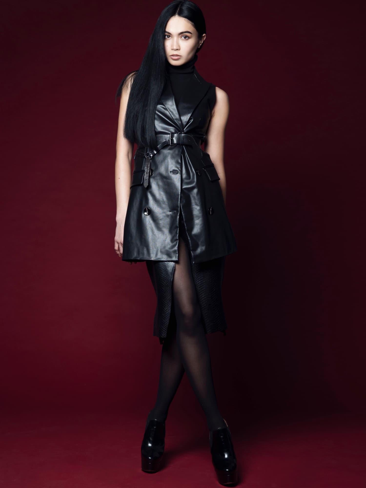 Shirin Hojayeva stands against a deep burgundy backdrop in a sleeveless leather trench dress, looking sleek and commanding.