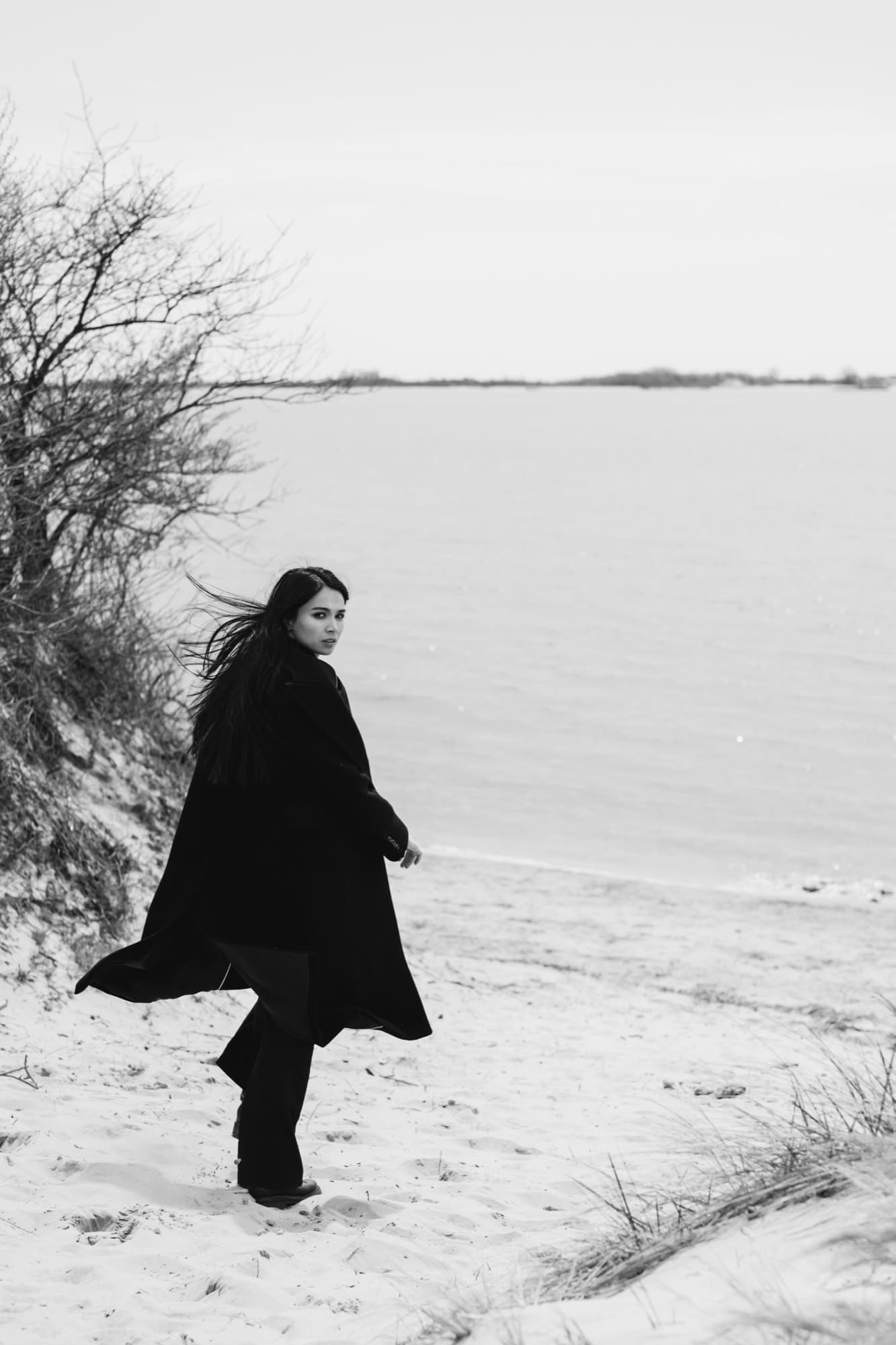 Shirin Hojayeva walks along a windswept winter beach in a long black coat, glancing back with a wistful expression.