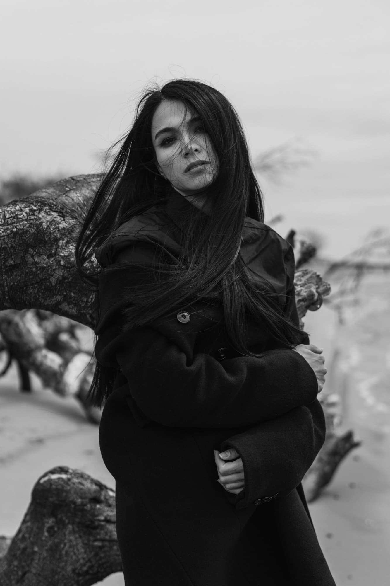 Shirin Hojayeva wraps herself in a dark coat beside driftwood on a beach, hair blowing across her face, looking moody.