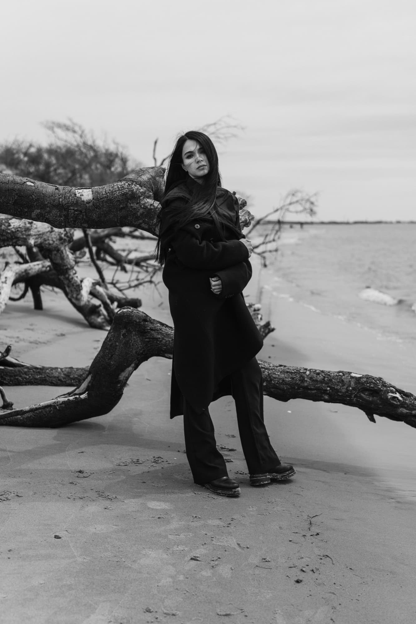Shirin Hojayeva leans against fallen tree branches on a beach in a black coat, conveying solitary strength.