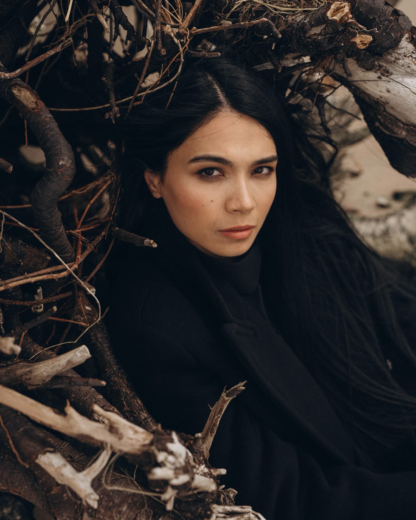 Close-up portrait of Shirin Hojayeva nestled among bare branches in a black coat, gazing up with raw vulnerability.