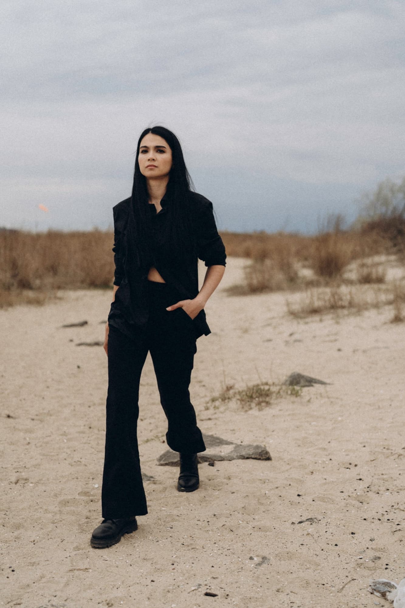 Shirin Hojayeva stands on a sandy shore in all black with hands in pockets, looking contemplative under an overcast sky.