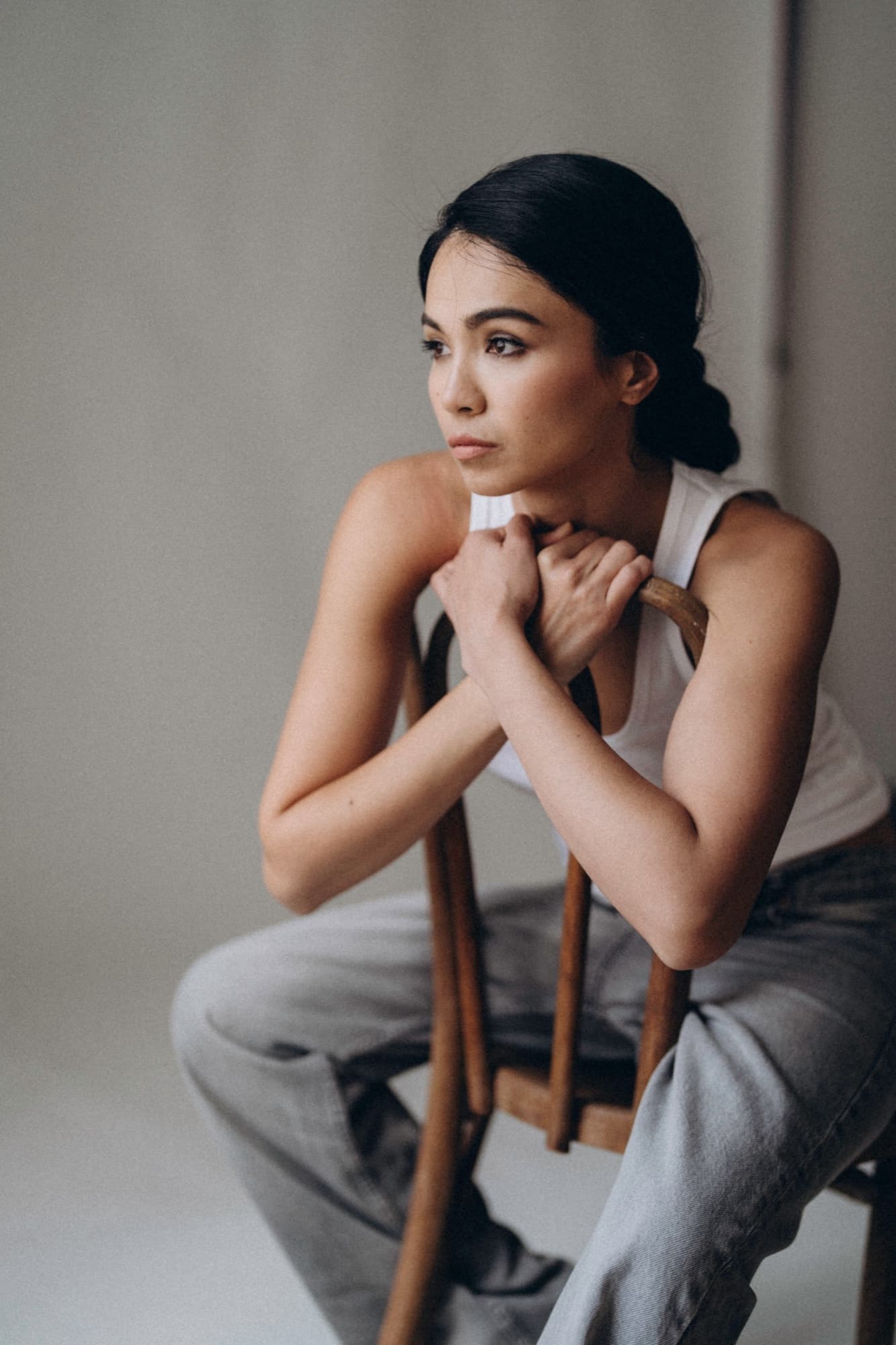 Shirin sits pensively on a wooden chair in a minimal studio, hands clasped, wearing a white tank top and jeans.