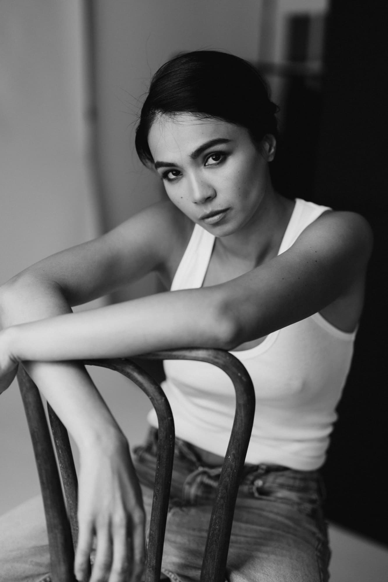 Black-and-white portrait of Shirin leaning on a bentwood chair in a white tank, gazing directly at the camera.