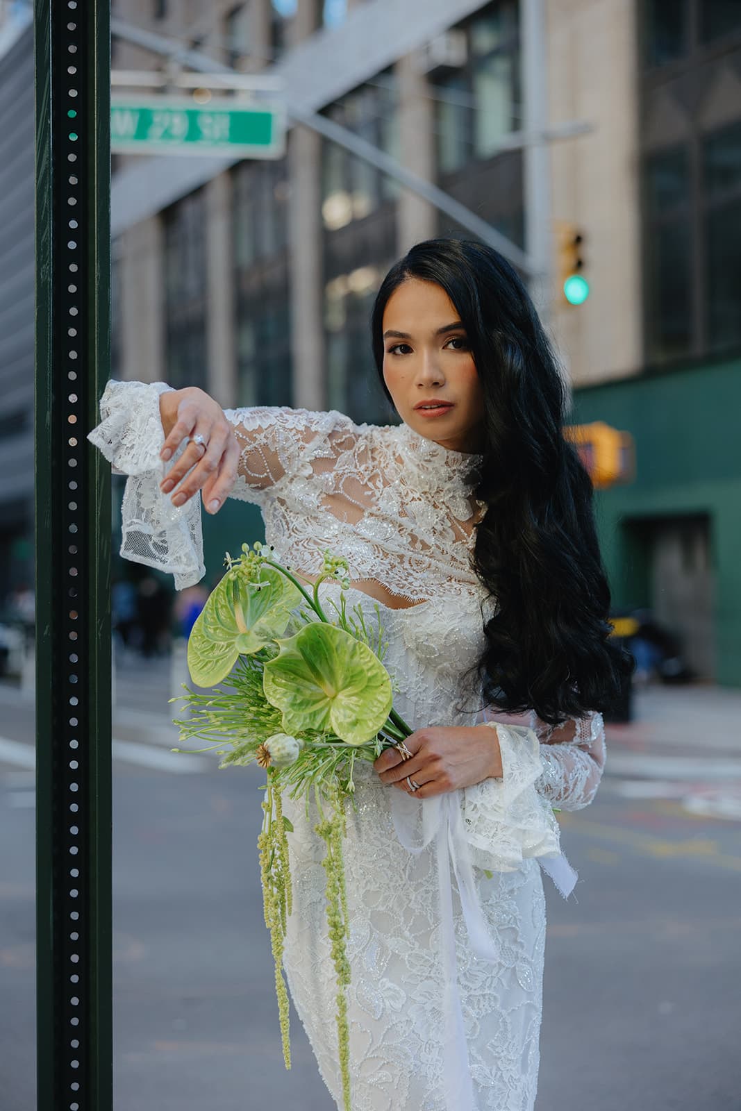 Shirin poses on a Manhattan street in a white lace bridal gown, holding a green anthurium bouquet with quiet poise.