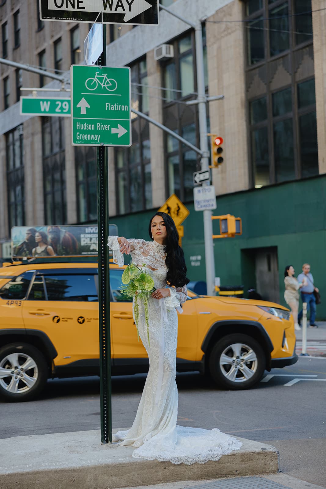 Shirin stands by a street sign in a white lace gown with bouquet as yellow cabs pass on a busy NYC block.