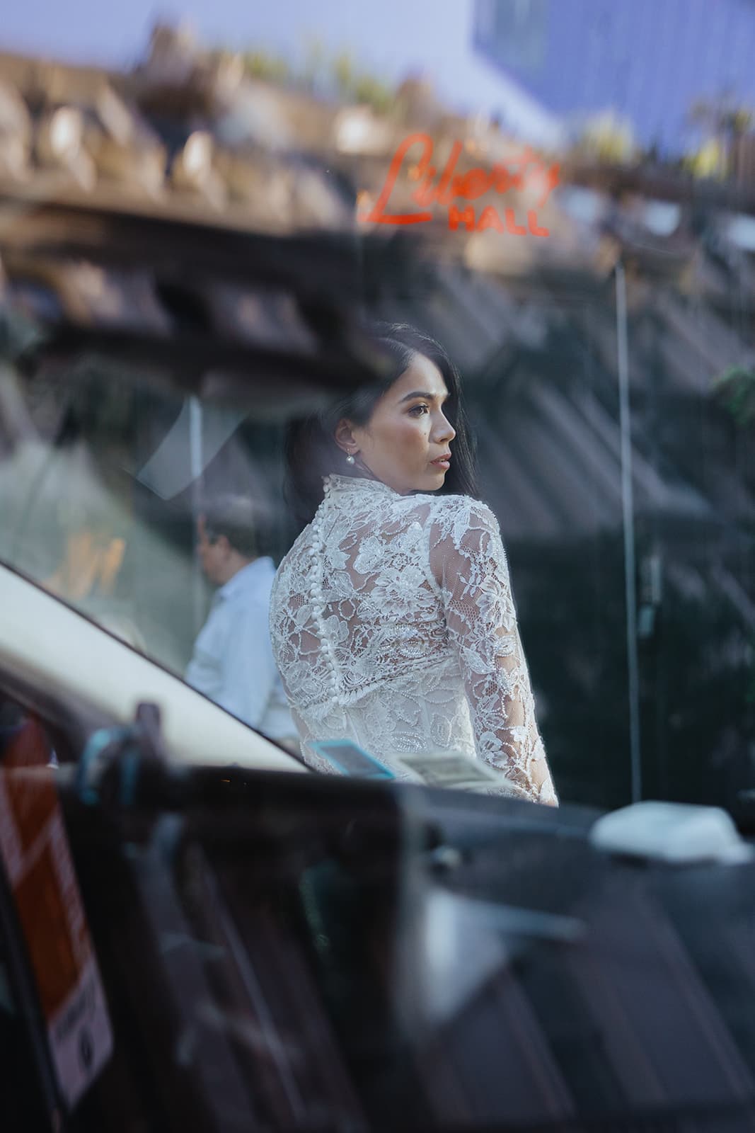 Shirin in a white lace dress seen through a window reflection, glancing over her shoulder with a contemplative gaze.