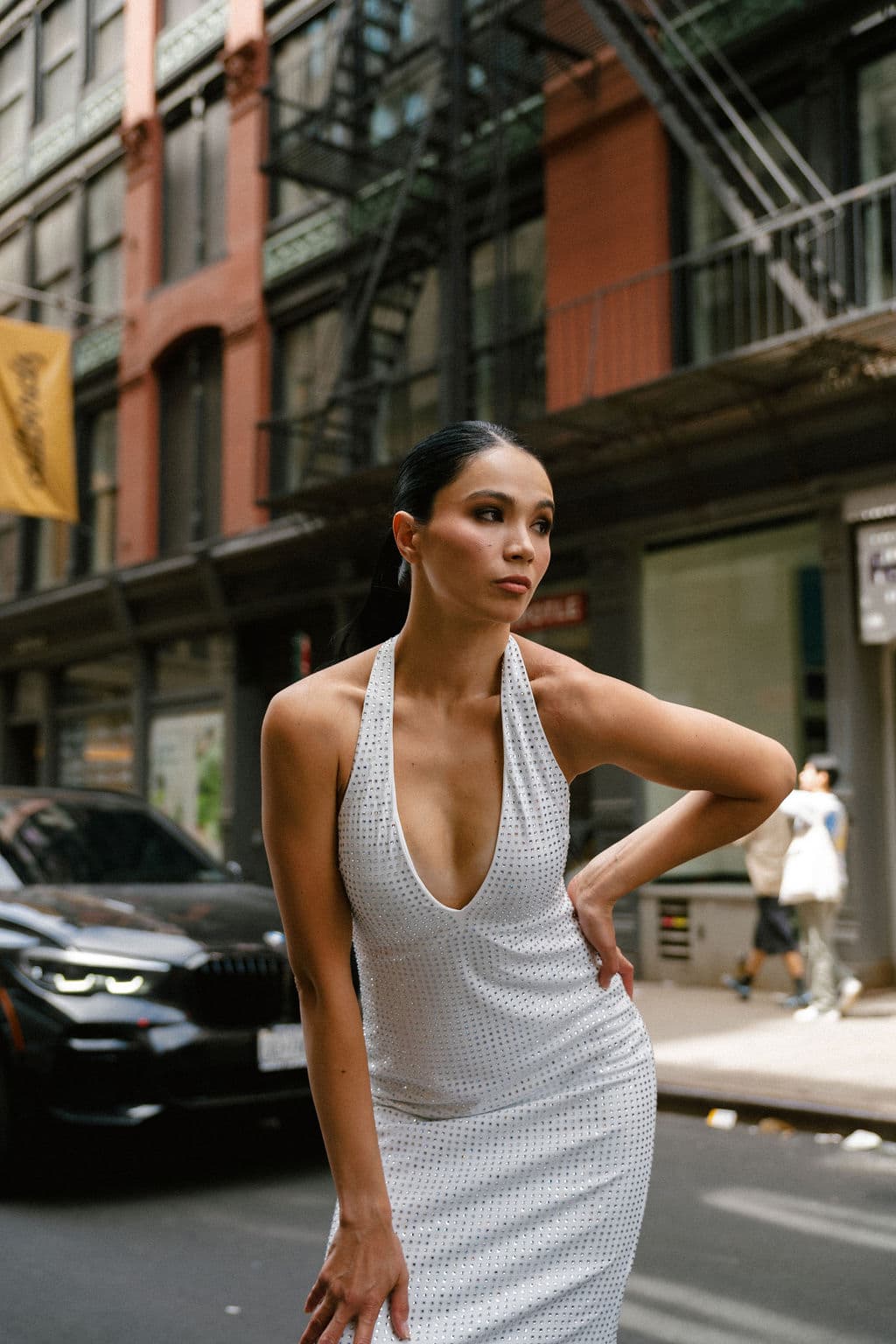 Shirin poses confidently on a SoHo street in a white studded halter dress, hand on hip, with a bold gaze.