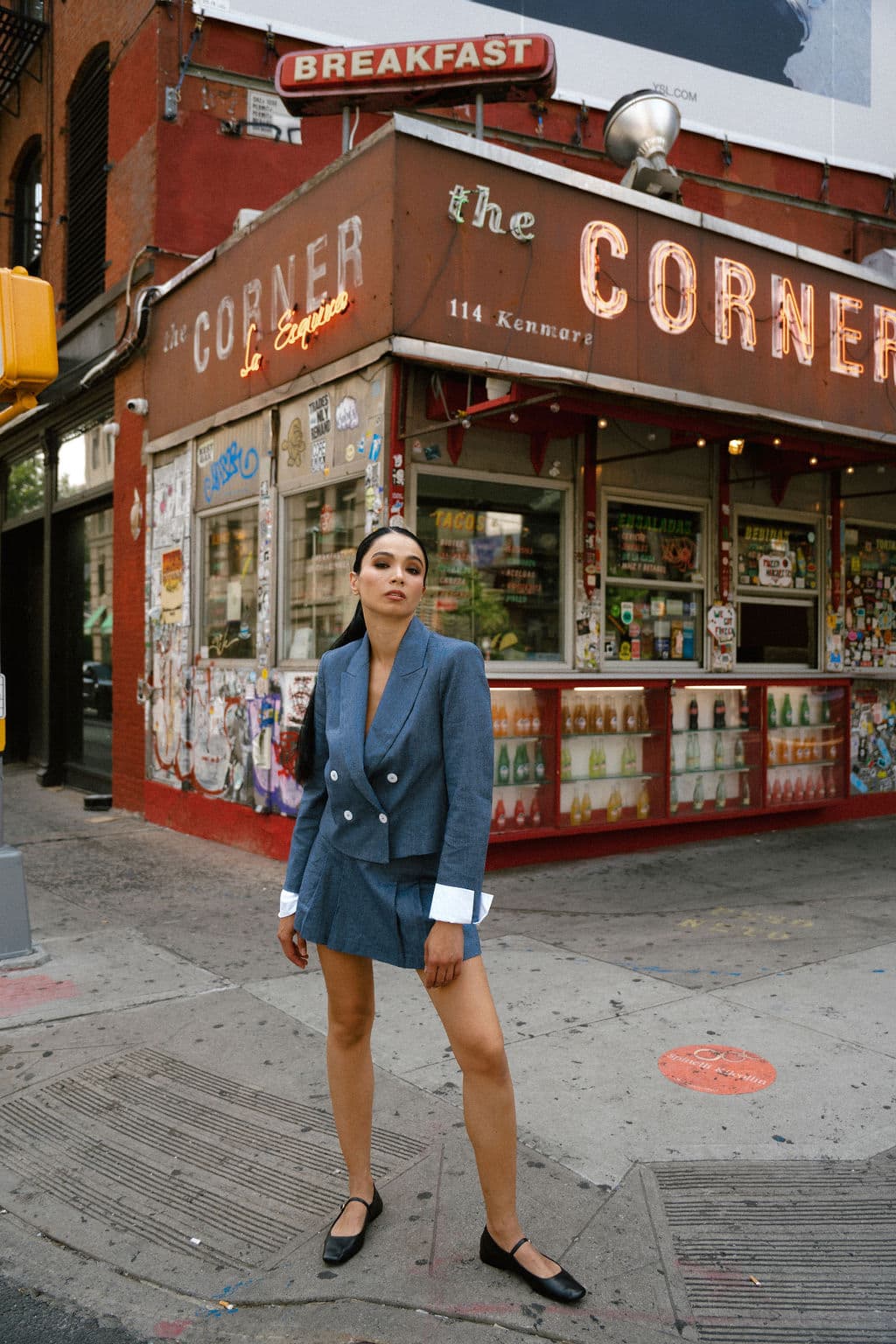 Shirin stands before a corner bodega in a blue double-breasted blazer and mini skirt, striking a poised stance.