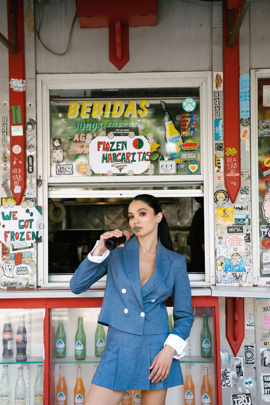 Shirin sips from a bottle outside a colorful bodega window in a blue blazer set, cool and self-assured.
