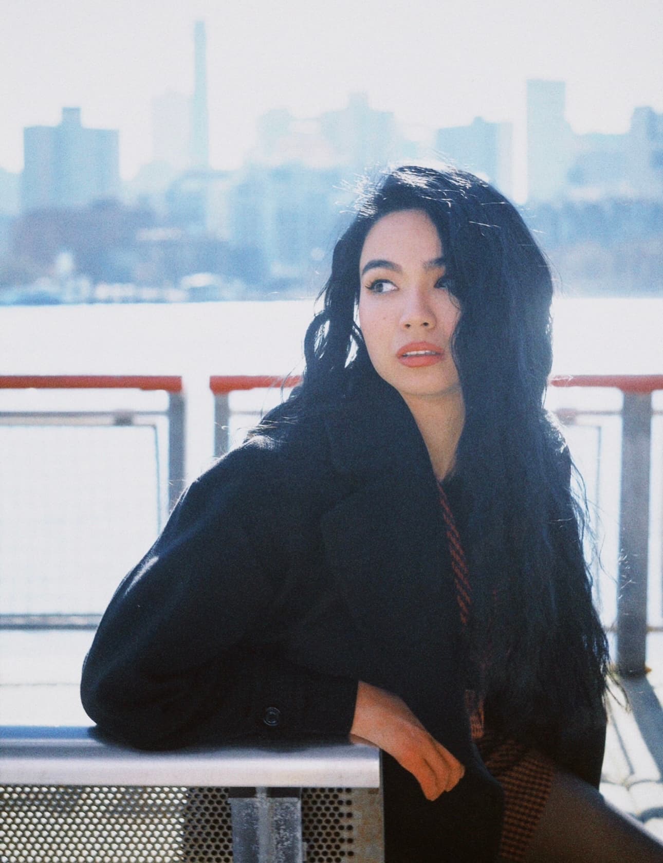 Shirin leans on a ferry railing in a dark coat, windswept hair framing her face against a hazy city skyline.