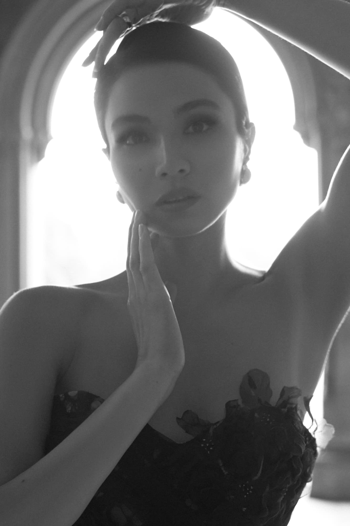 Black-and-white close-up of Shirin backlit in an archway, hand near her chin, wearing a strapless floral top.