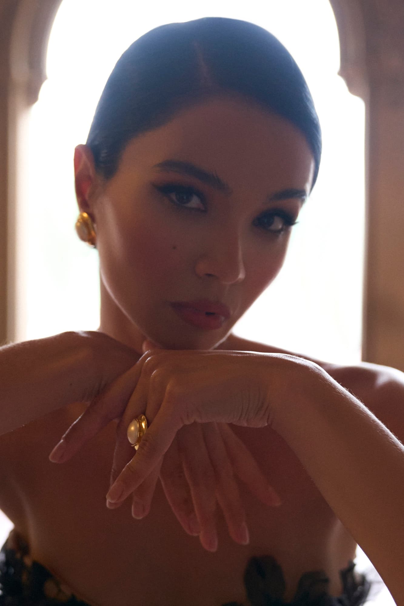 Tight portrait of Shirin with slicked-back hair and gold jewelry, hands crossed at her chest in warm golden light.