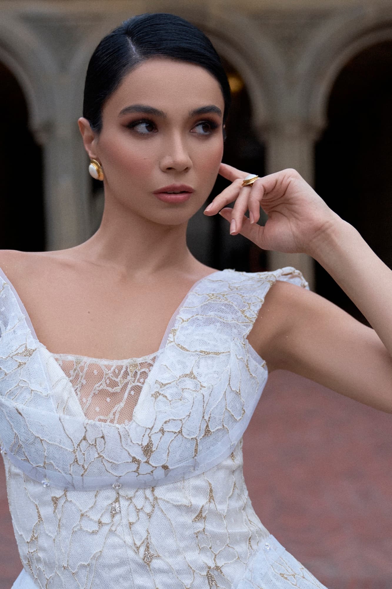Close-up of Shirin in a white lace gown with gold earrings, delicately touching her cheek at Bethesda Terrace.