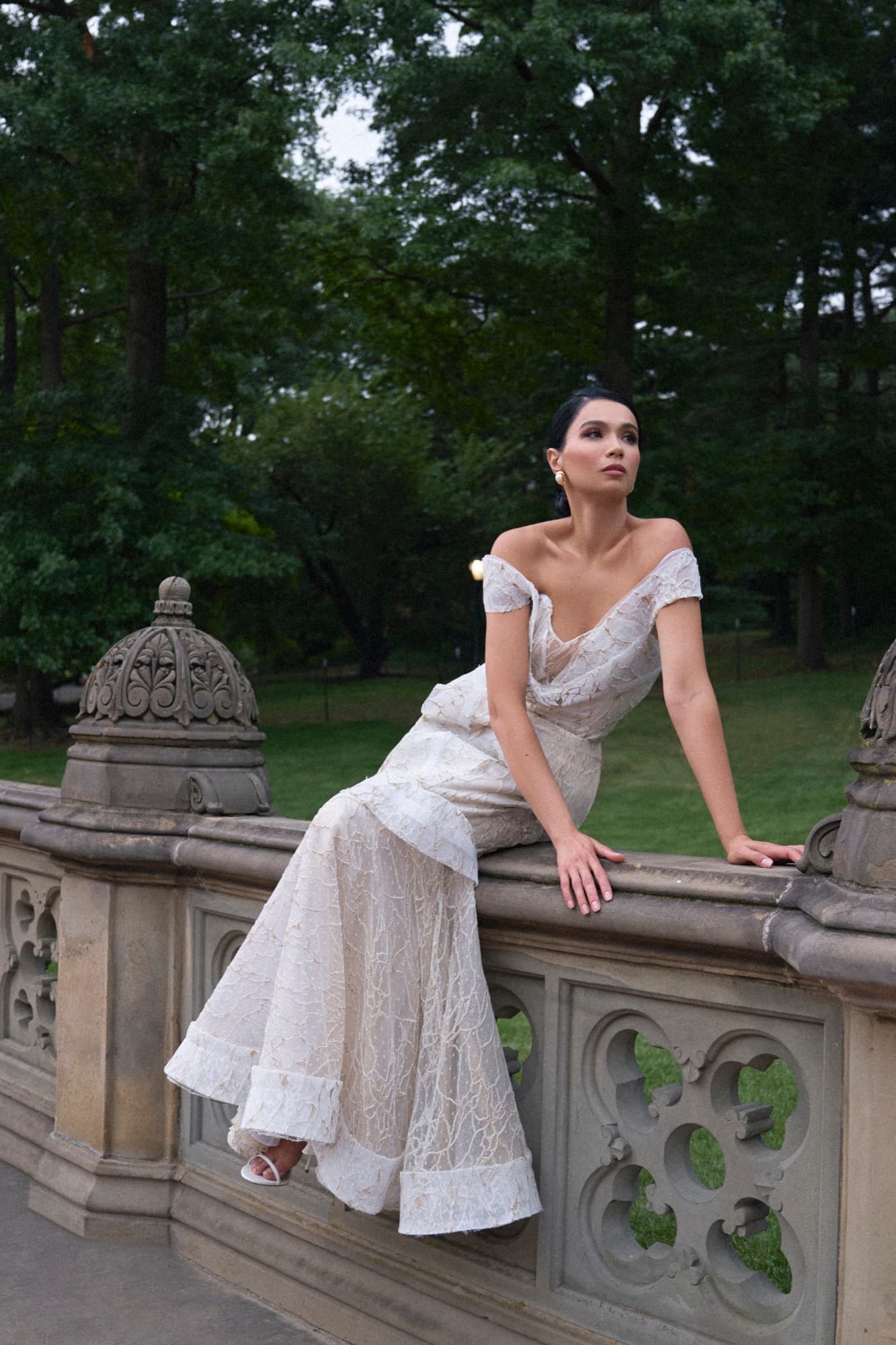 Shirin sits on a stone balustrade in Central Park wearing an off-shoulder lace gown, gazing upward serenely.