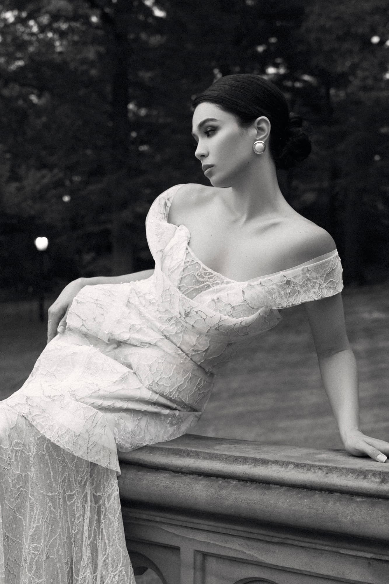 Black-and-white portrait of Shirin seated on a stone railing in a lace gown, looking down with quiet grace.