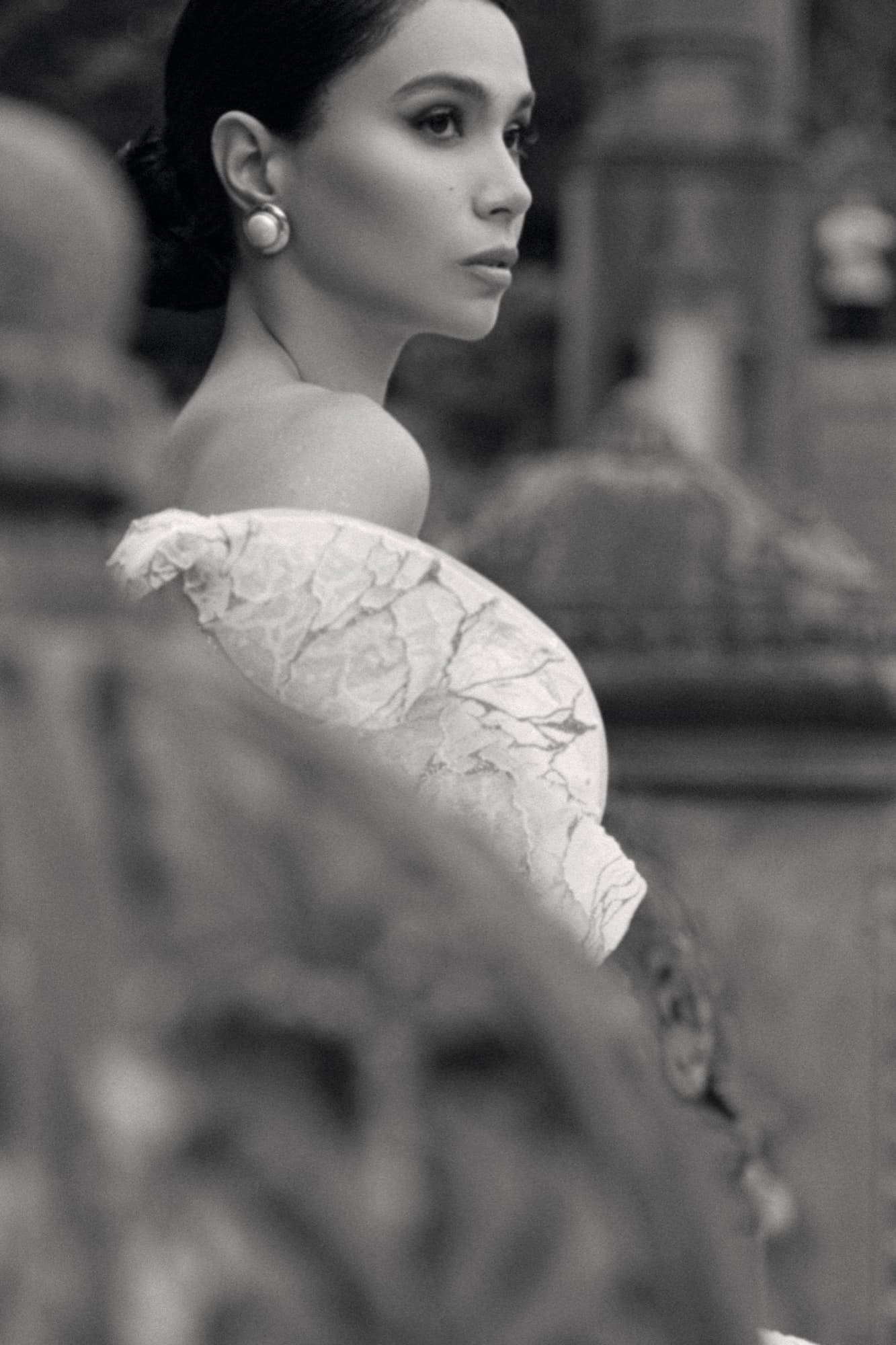 Black-and-white over-the-shoulder portrait of Shirin in a lace gown and pearl earring, framed by soft bokeh.