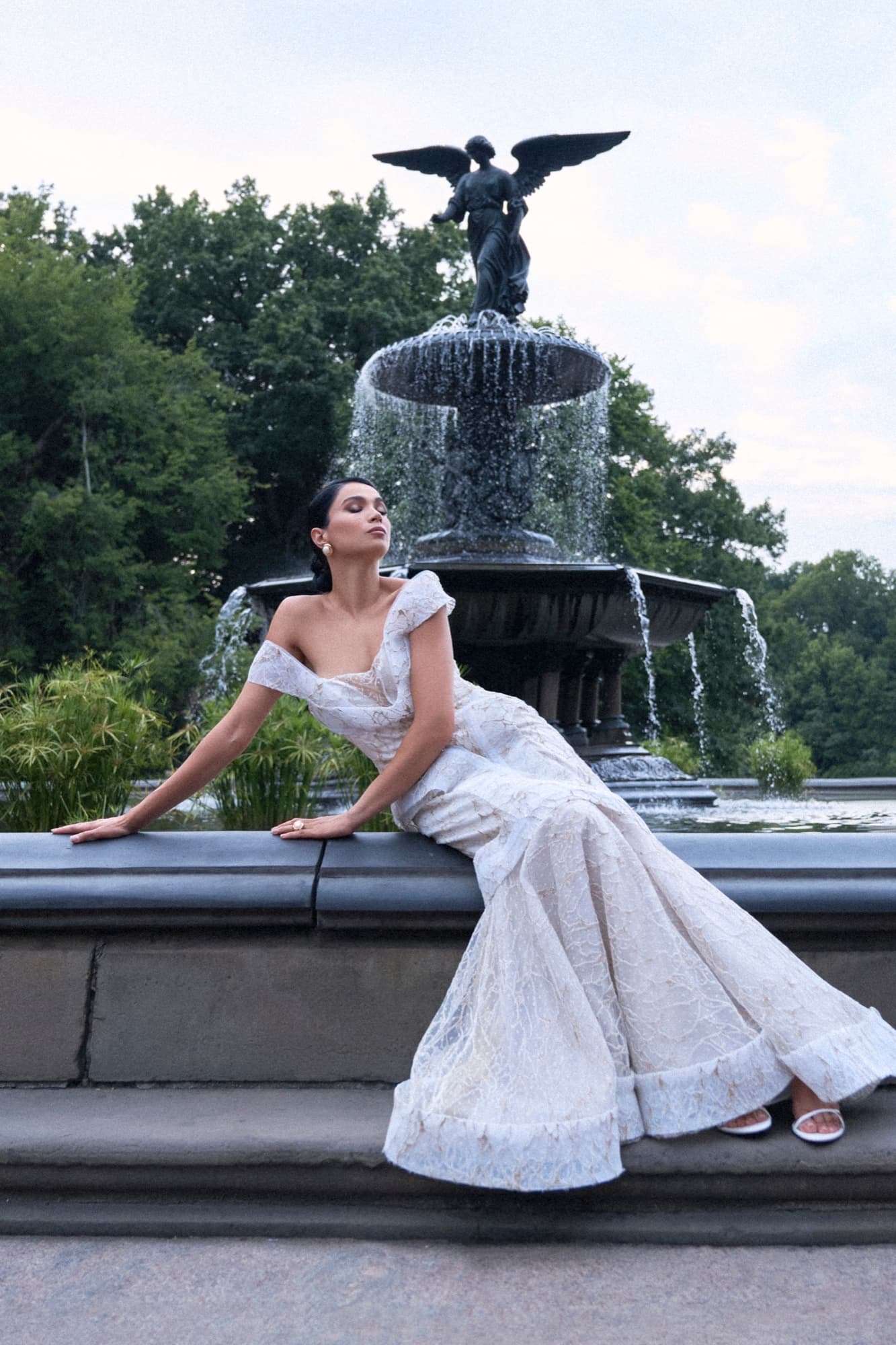 Shirin reclines on the Bethesda Fountain ledge in a white lace gown, eyes closed, with the angel statue behind her.