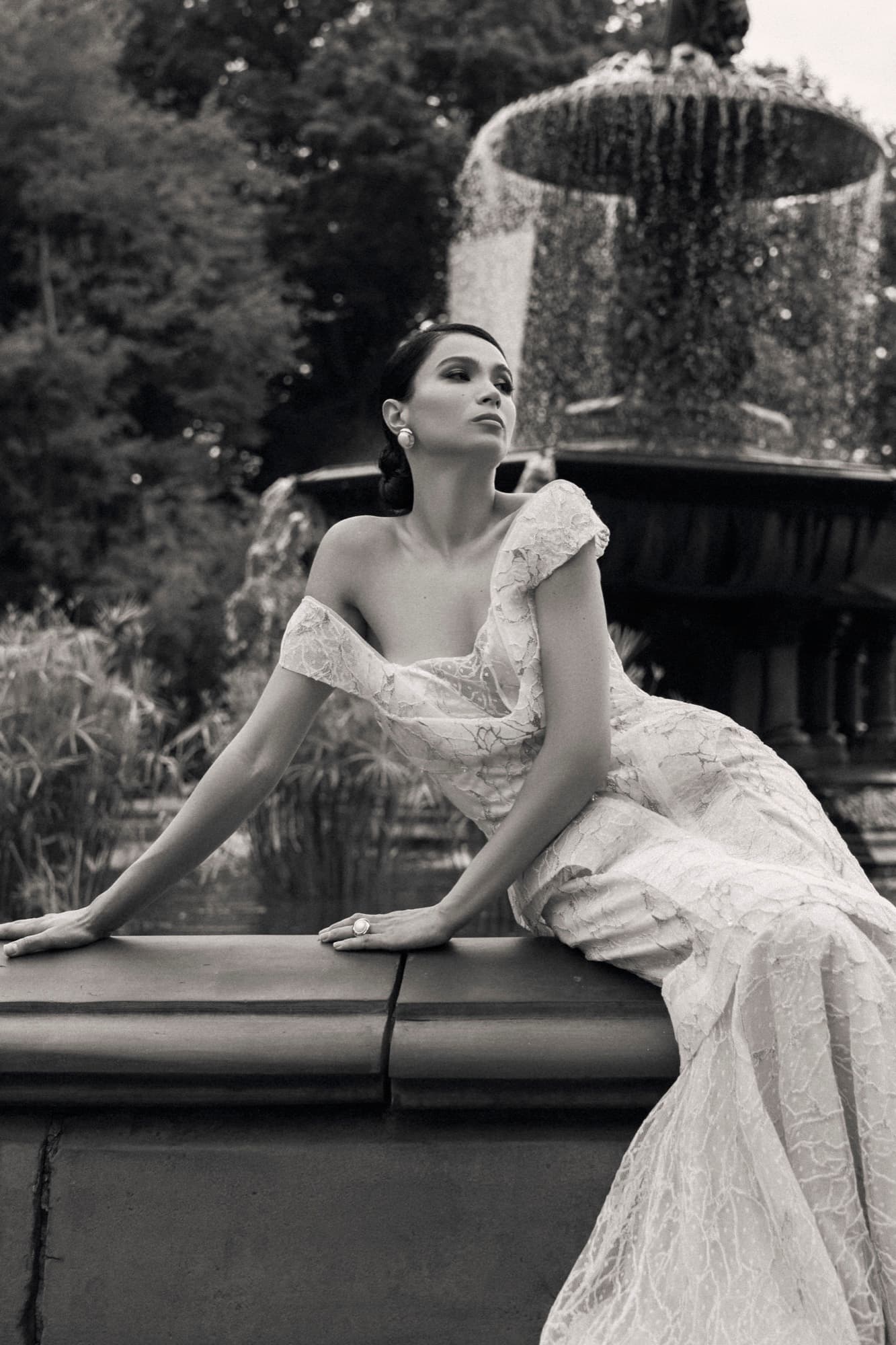 Shirin in a lace off-shoulder gown leans against a park fountain, gazing upward in a timeless black-and-white portrait.