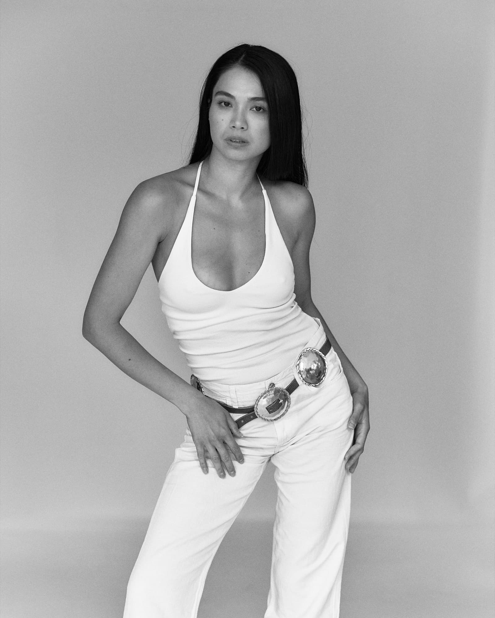 Shirin stands in a white halter top and jeans with a concho belt against a studio backdrop, exuding casual confidence.