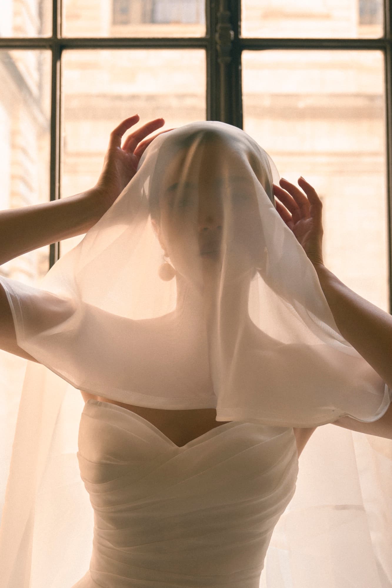 Shirin lifts a sheer veil over her face by a sunlit window in a strapless white gown, evoking dreamy mystery.