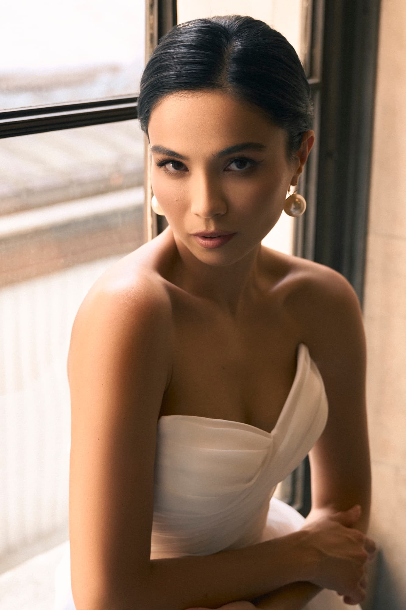 Shirin leans by a window in a strapless white gown with pearl earrings, giving a warm and intimate gaze.