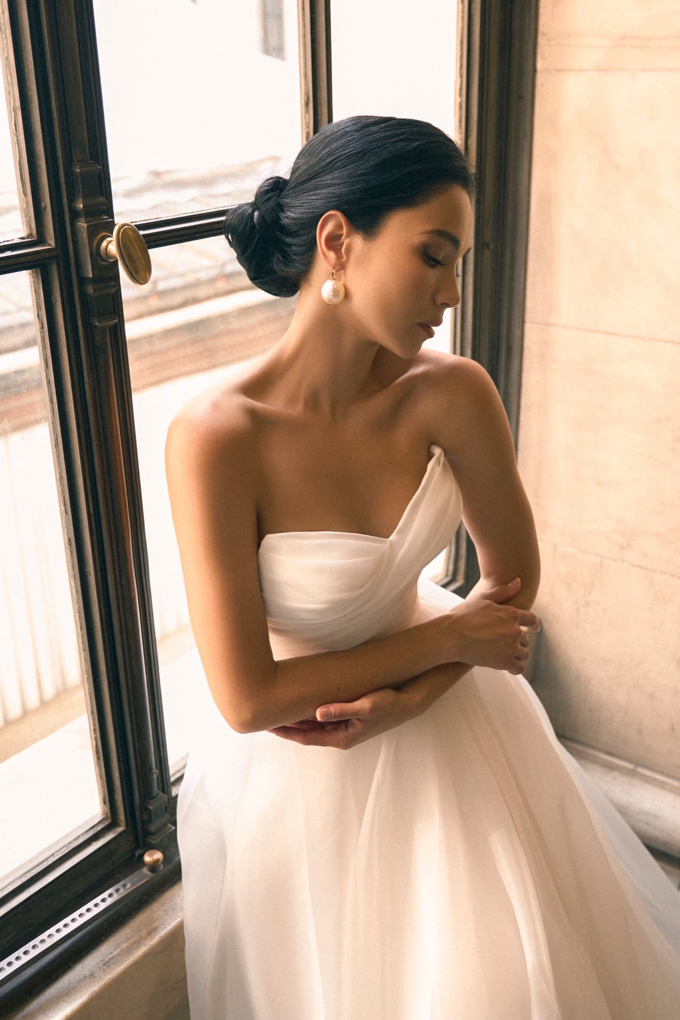 Shirin stands by a tall window in a strapless tulle gown, eyes downcast with hands clasped, quietly contemplative.