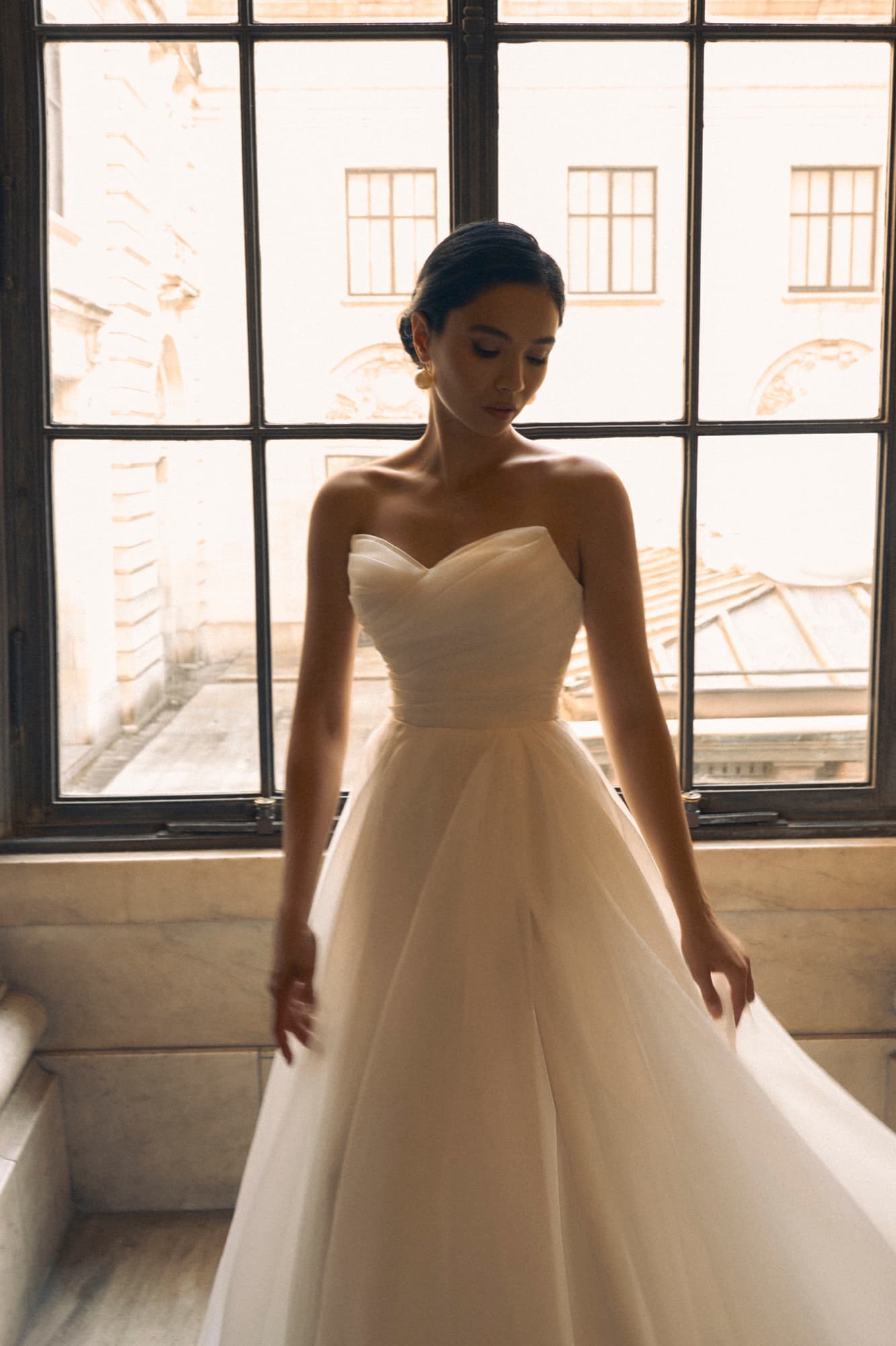 Shirin in a sweetheart tulle bridal gown stands before a grand window, looking down softly as the skirt cascades.