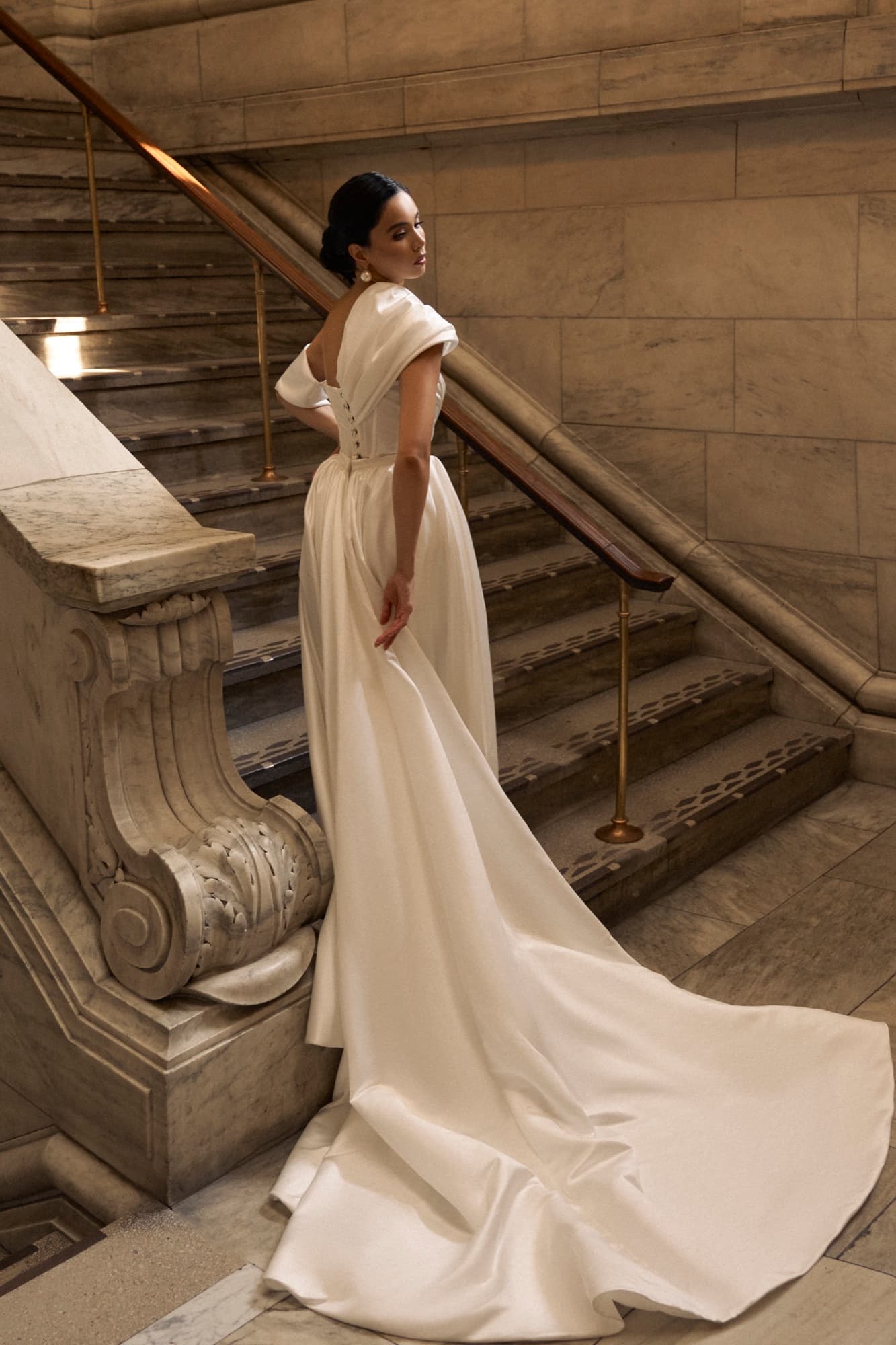 Shirin ascends a grand marble staircase in a satin gown with a long train, glancing back with quiet poise.