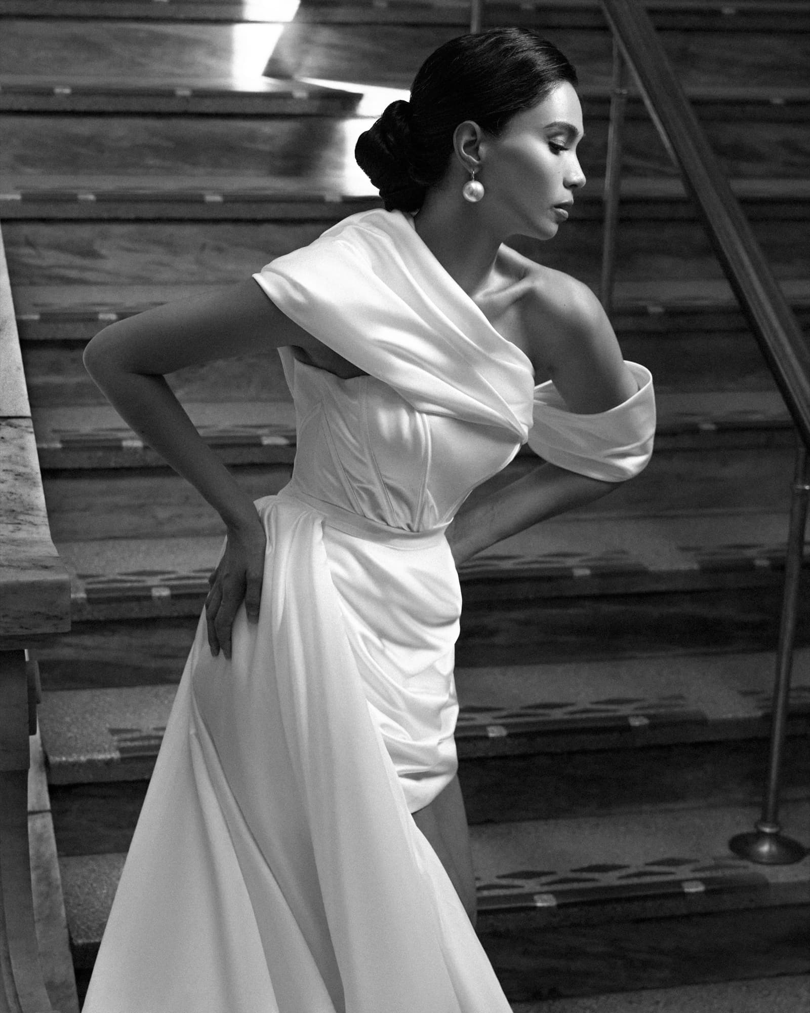 Shirin poses on grand stairs in a draped off-shoulder satin gown with pearl earring, hand on hip, looking away.