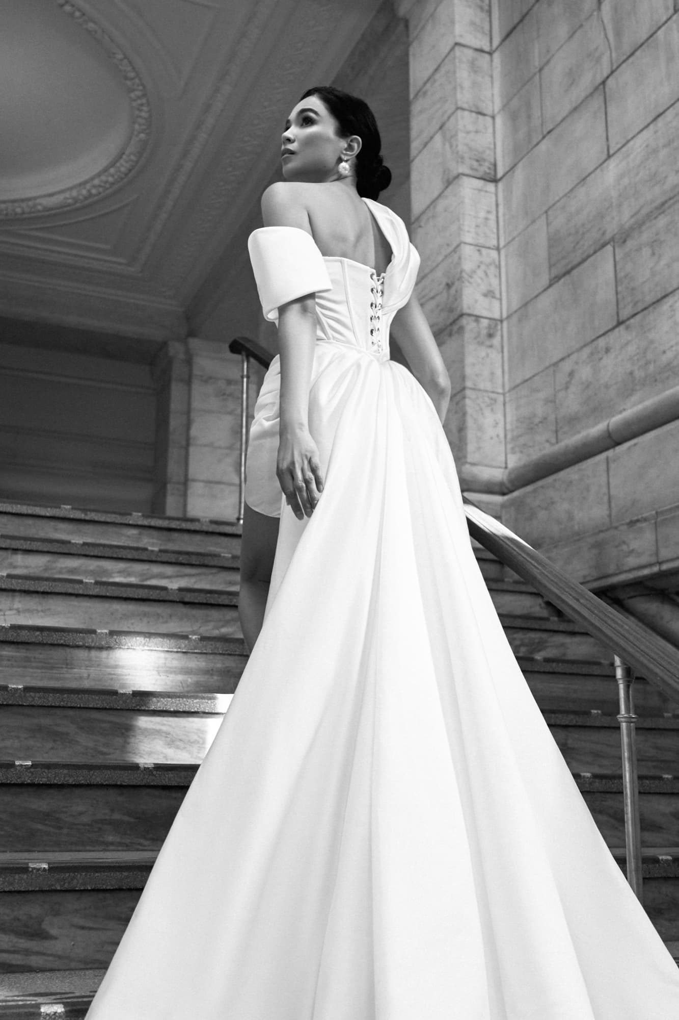 Black-and-white low-angle shot of Shirin ascending marble stairs in an off-shoulder corset gown with a sweeping train.
