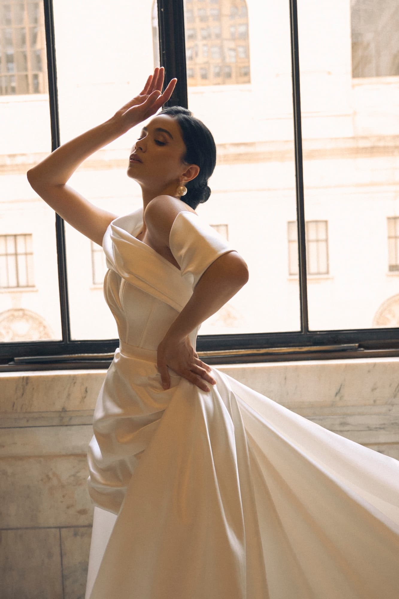 Shirin raises one hand gracefully by a sunlit window in an off-shoulder satin gown, eyes closed in elegant motion.