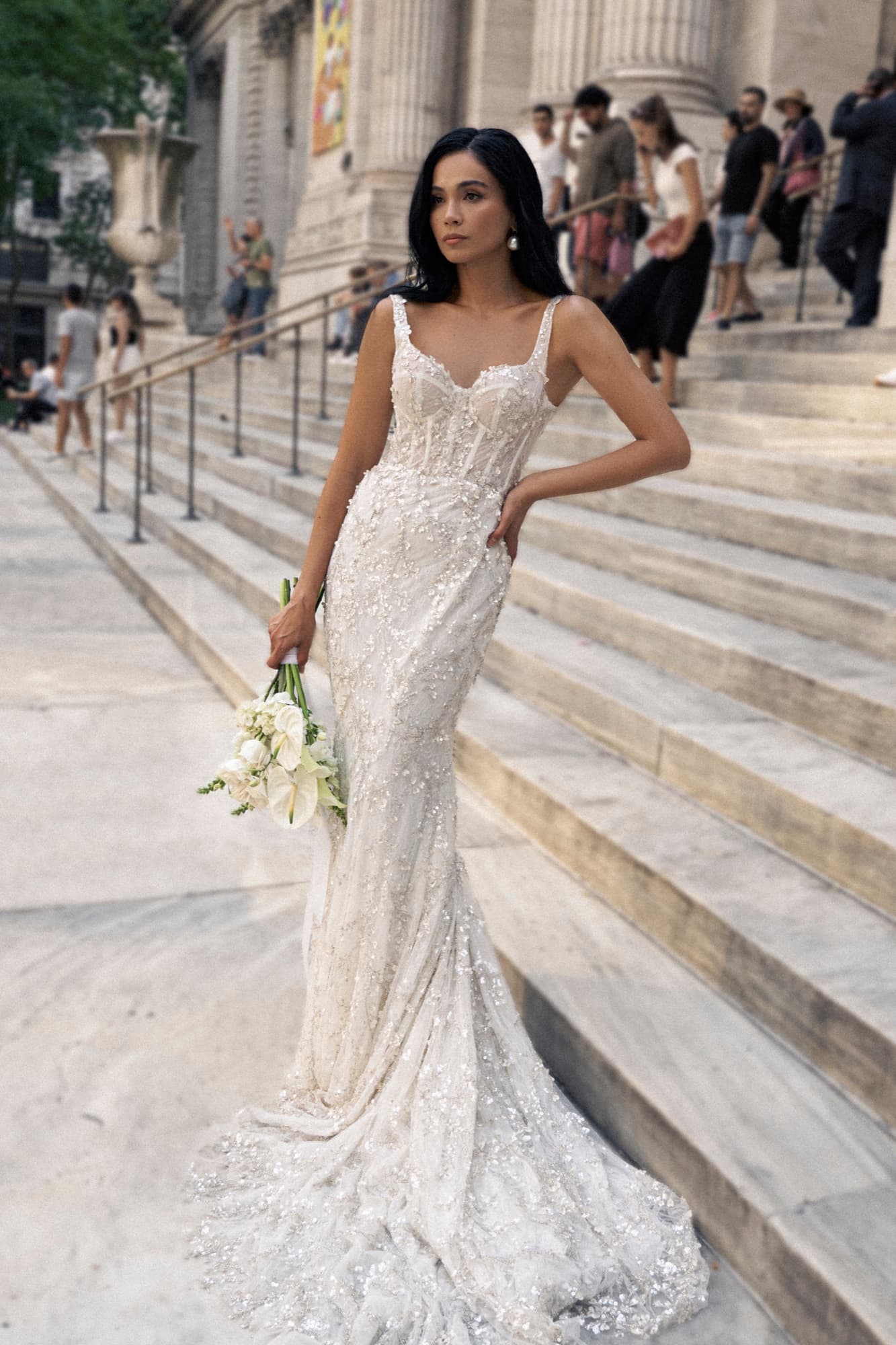 Shirin stands on stone steps in a beaded bridal gown holding a white bouquet, looking poised amid a city backdrop.