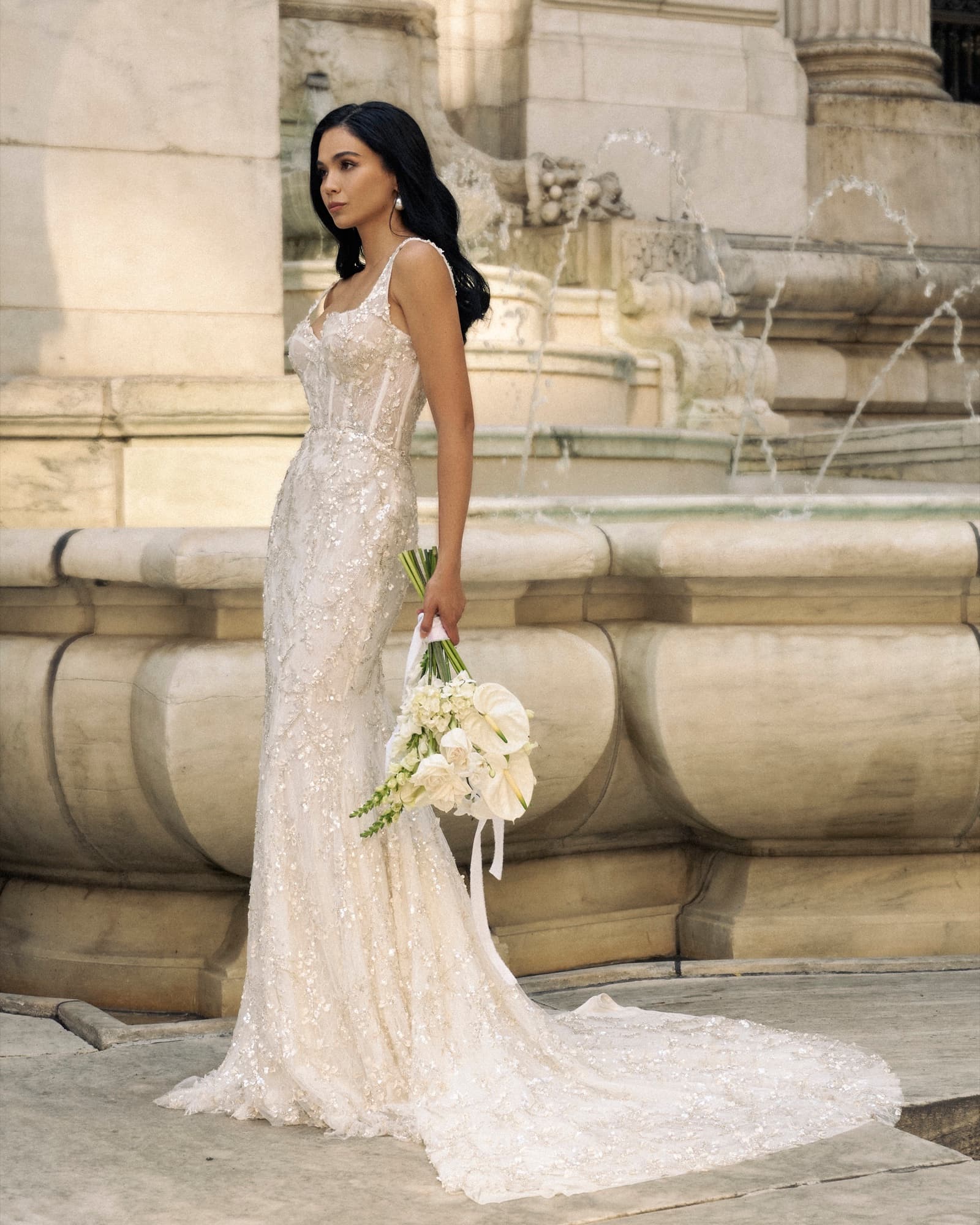 Shirin in a beaded gown with long train stands by a stone fountain holding a white bouquet, gazing into the distance.
