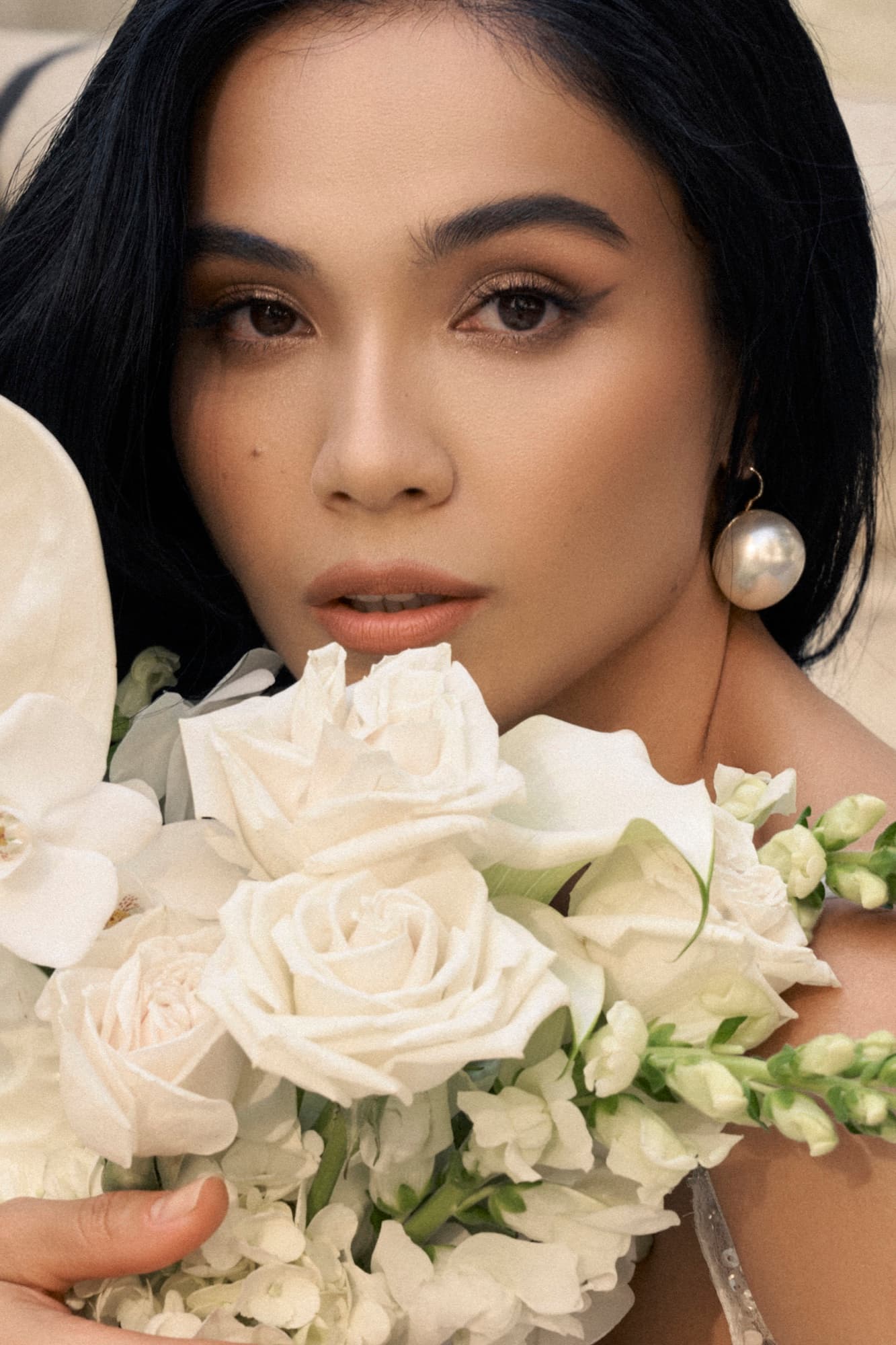 Close-up of Shirin resting her face near white roses and orchids, pearl earring visible, with a soft direct gaze.