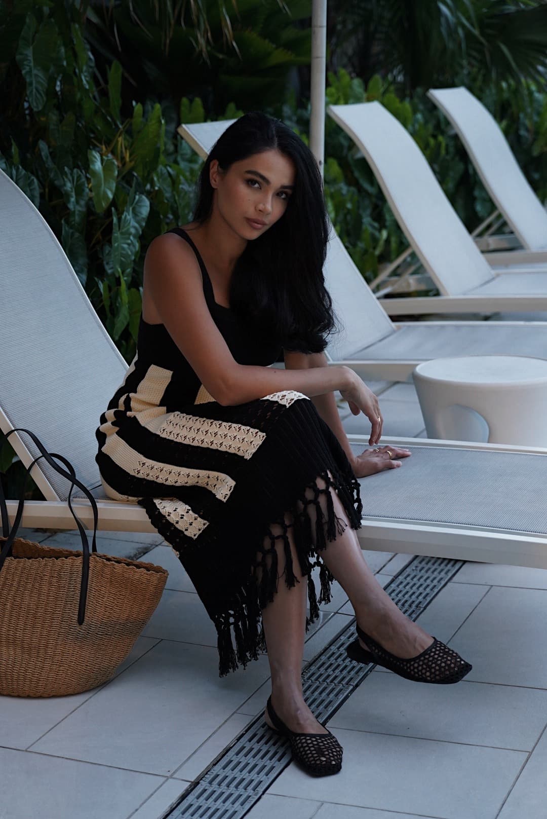 Shirin sits on a poolside lounge chair in a black-and-tan fringed knit dress, looking relaxed and effortlessly chic.
