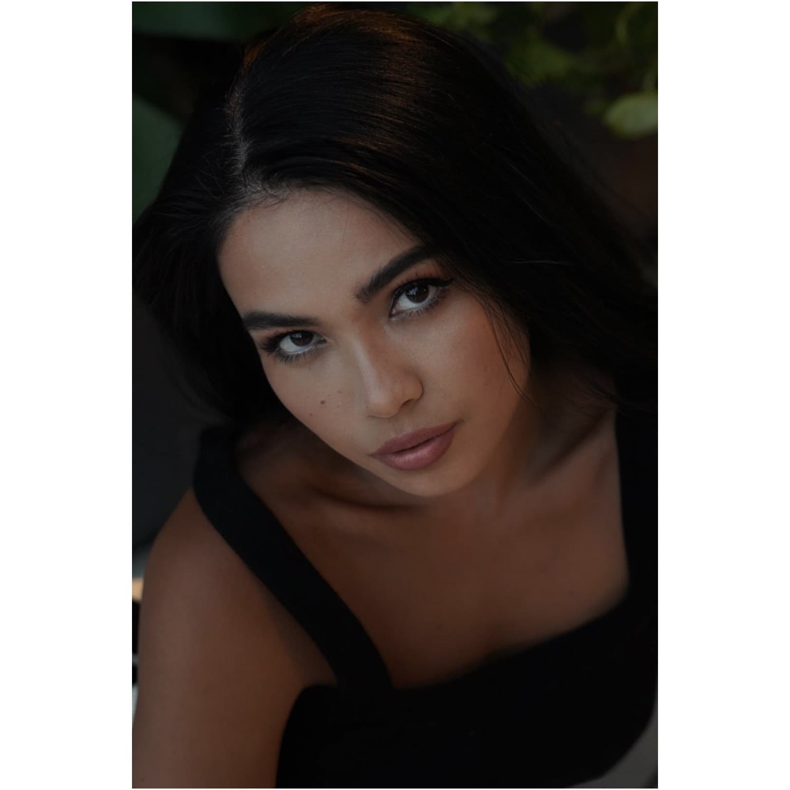 Overhead close-up of Shirin in a black top among dark foliage, gazing up at the camera with striking intensity.