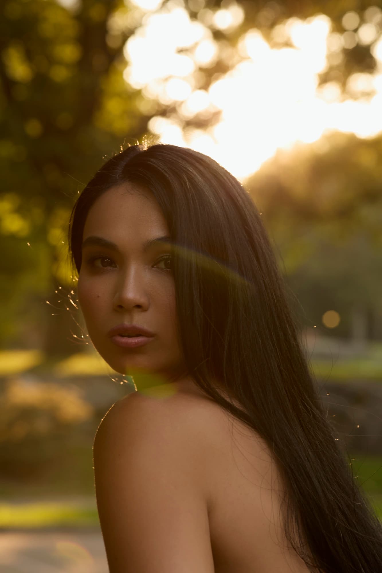 Shirin glances over her bare shoulder outdoors at golden hour, sun flare catching her flowing hair in a warm glow.