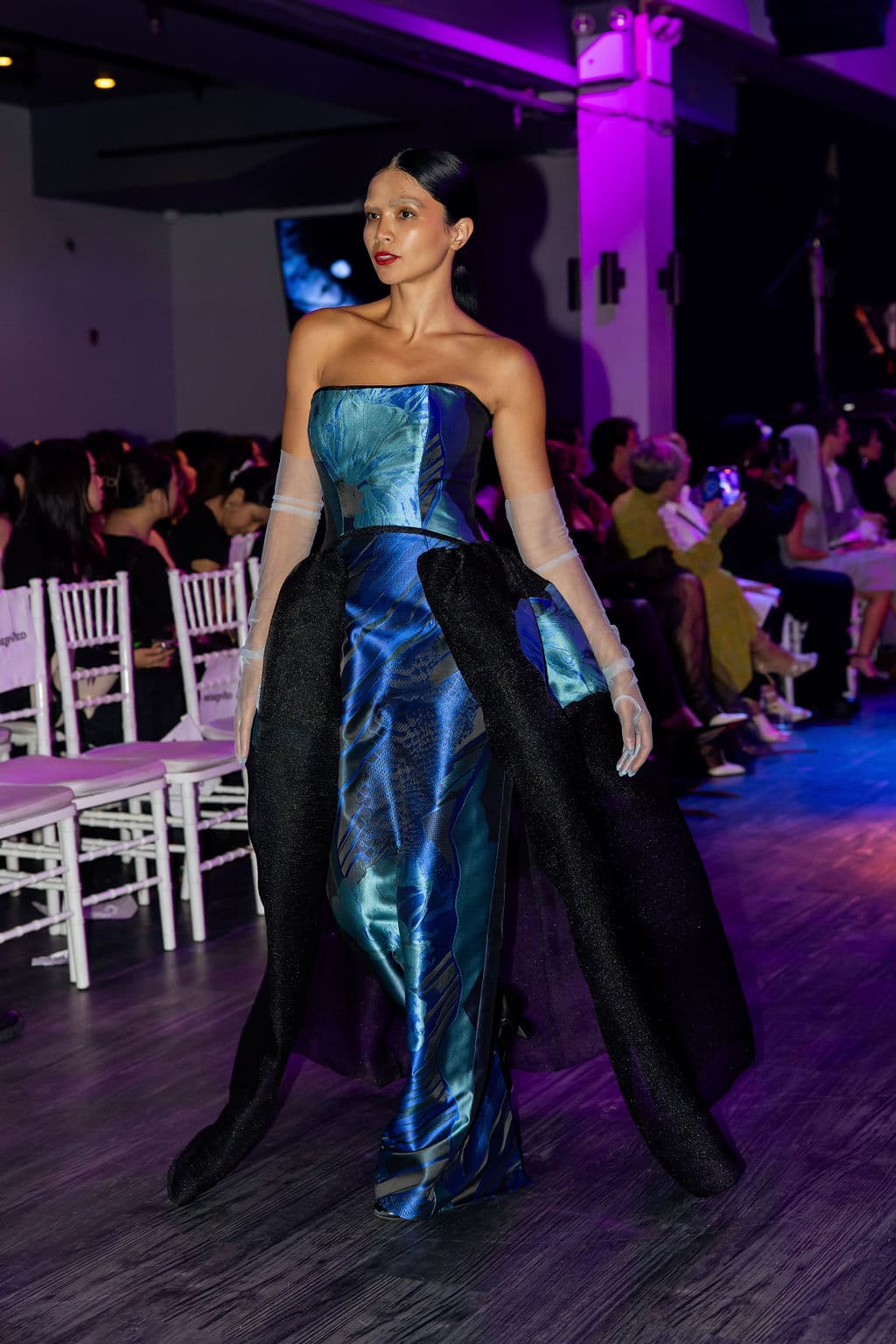 Shirin walks the runway in a strapless blue-and-black gown with sheer gloves under dramatic purple stage lighting.
