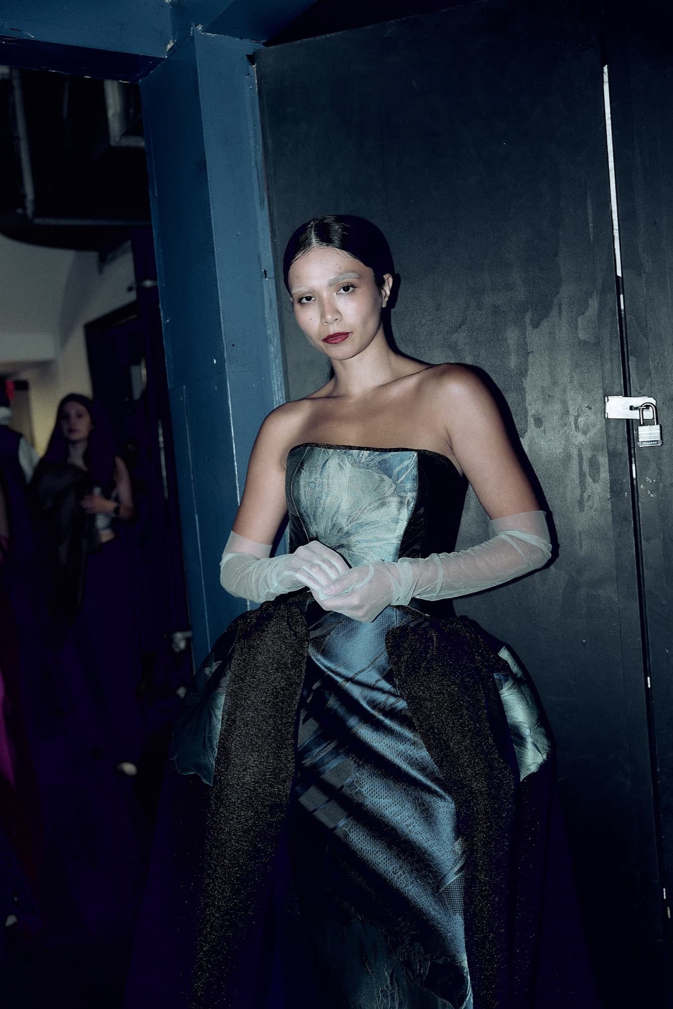 Shirin poses backstage in a sculptural blue-and-black strapless gown with sheer gloves, composed and runway-ready.