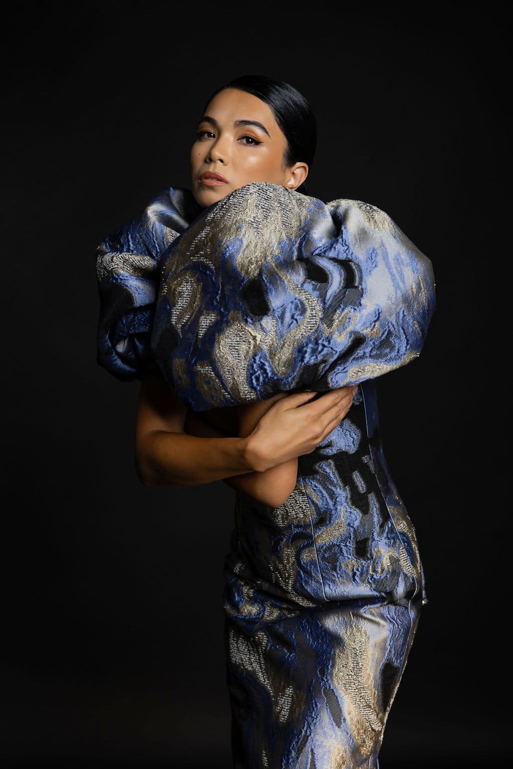 Shirin wraps herself in a blue-and-gold brocade puff-sleeve gown against a dark backdrop, gazing over the fabric.