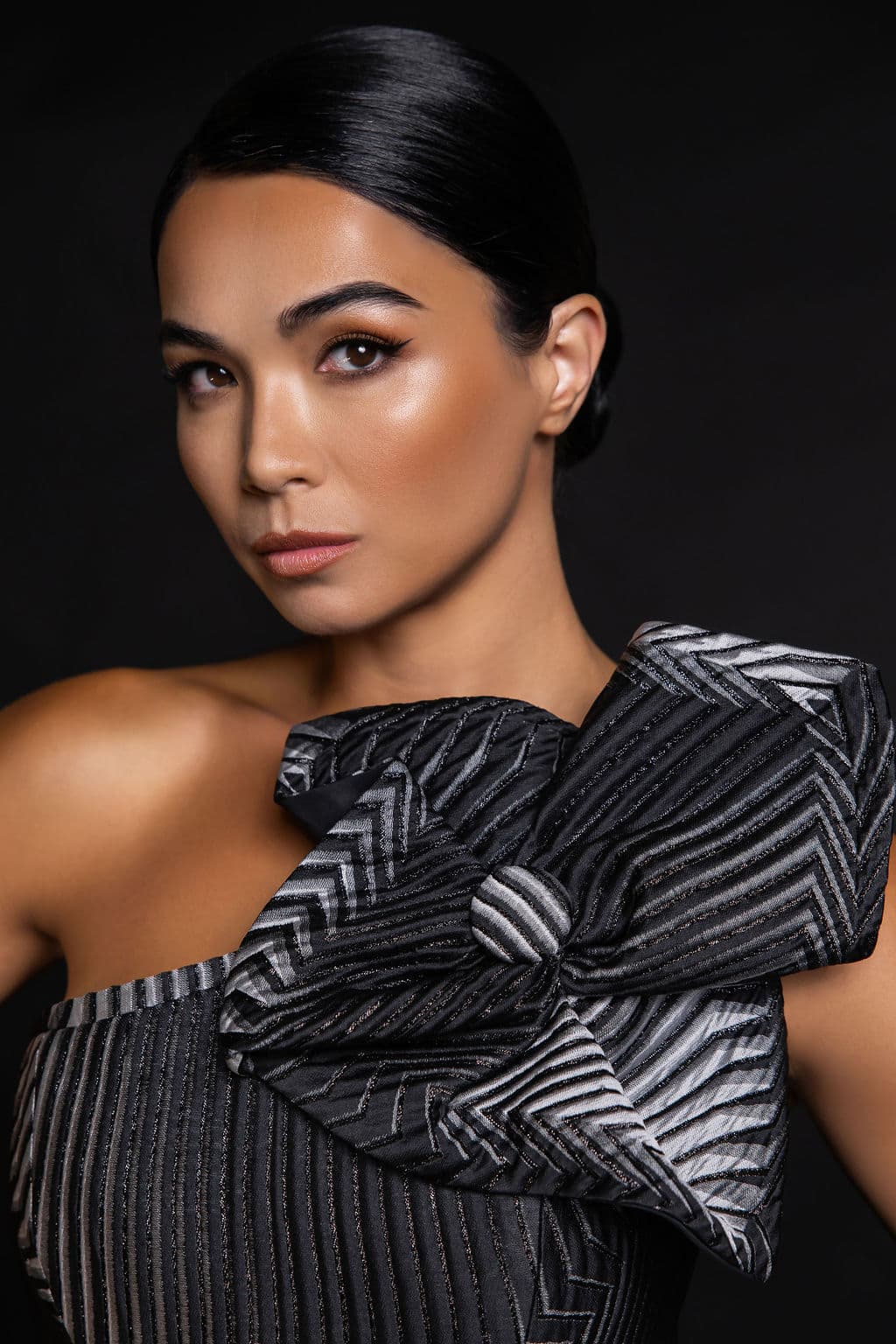 Close-up of Shirin Hojayeva in a sculptural black-and-silver striped bow-detail top against a dark backdrop, gaze intense.