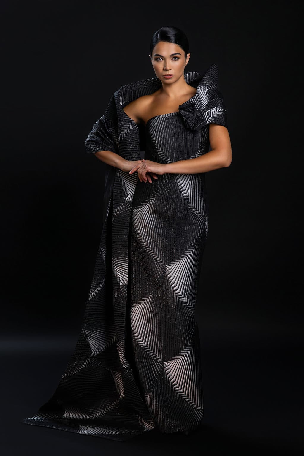 Shirin Hojayeva poses with clasped hands in a silver-striped geometric gown with dramatic shoulders against a black background.