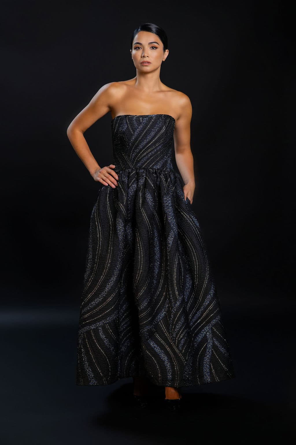 Shirin Hojayeva stands confidently in a dark textured strapless ball gown with gold wave details on a black backdrop.