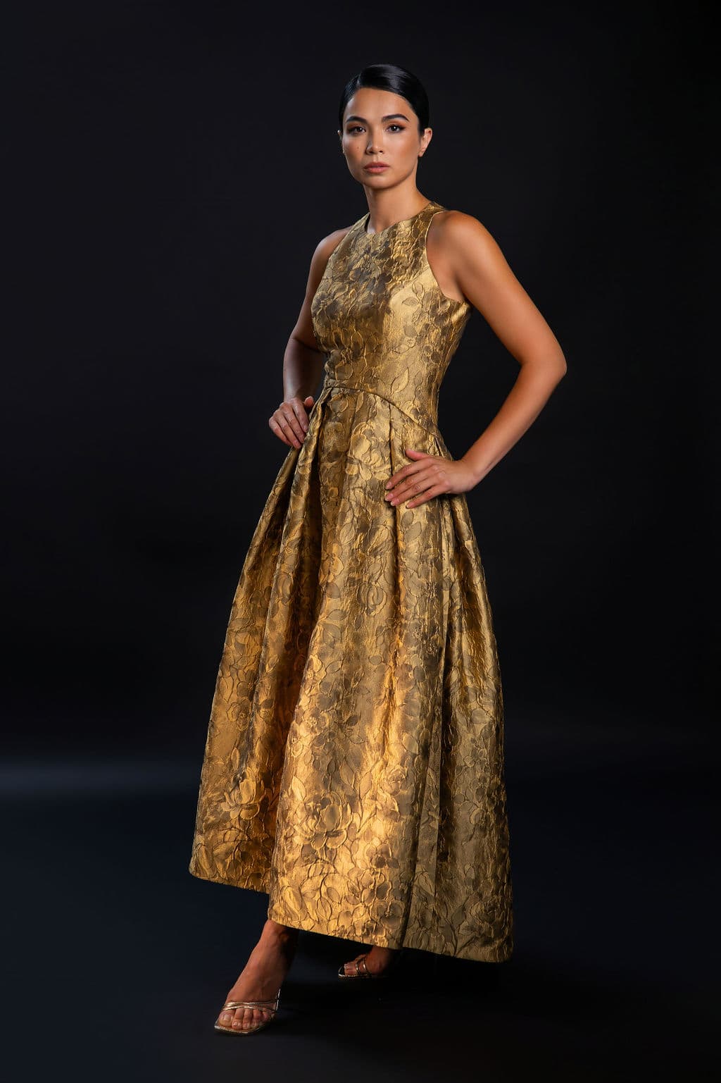Shirin Hojayeva poses in a gold floral brocade halter ball gown with hand on hip against a dark studio background.
