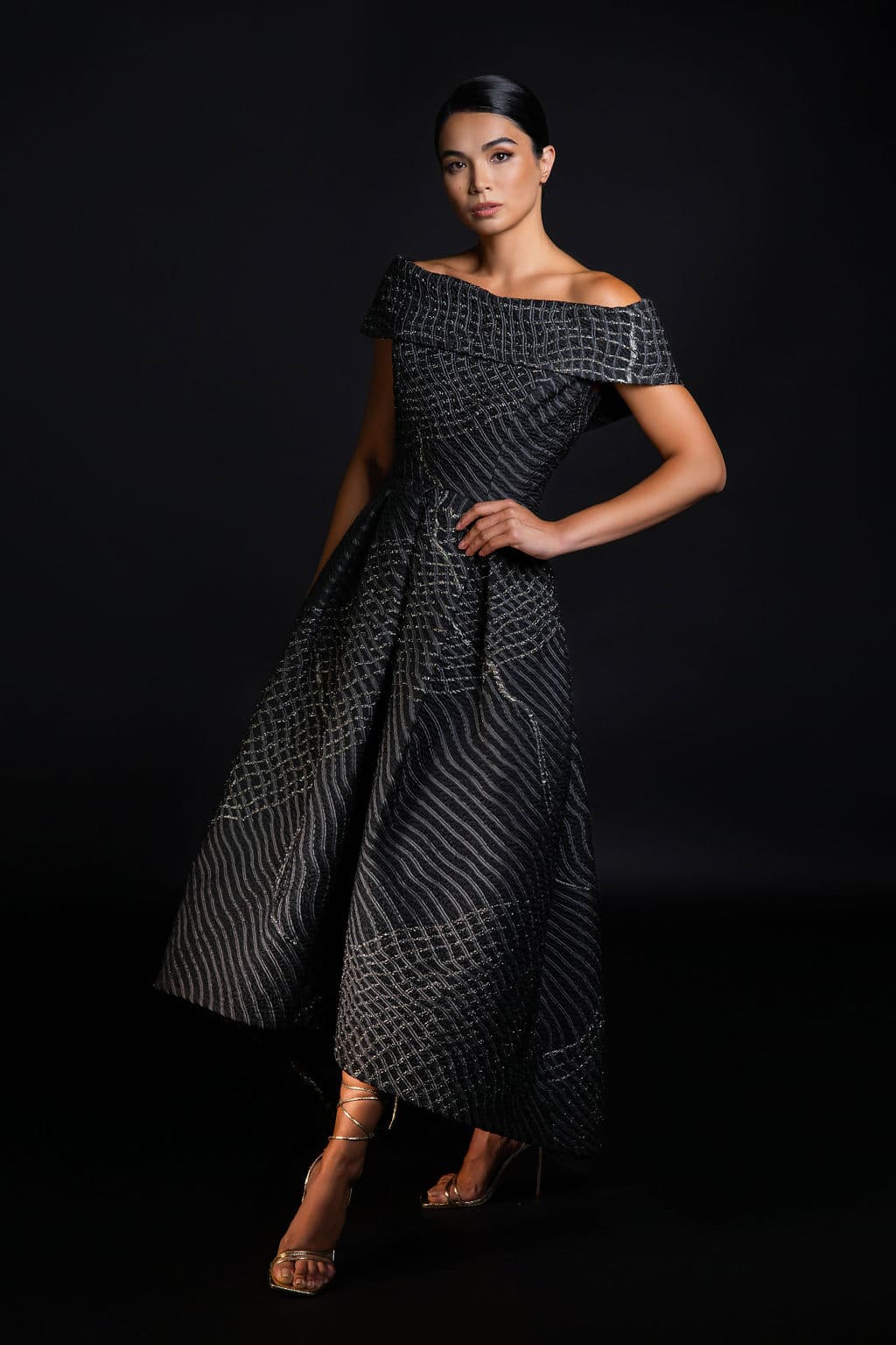 Shirin Hojayeva steps forward in a charcoal off-shoulder gown with geometric line patterns, poised and refined in studio light.