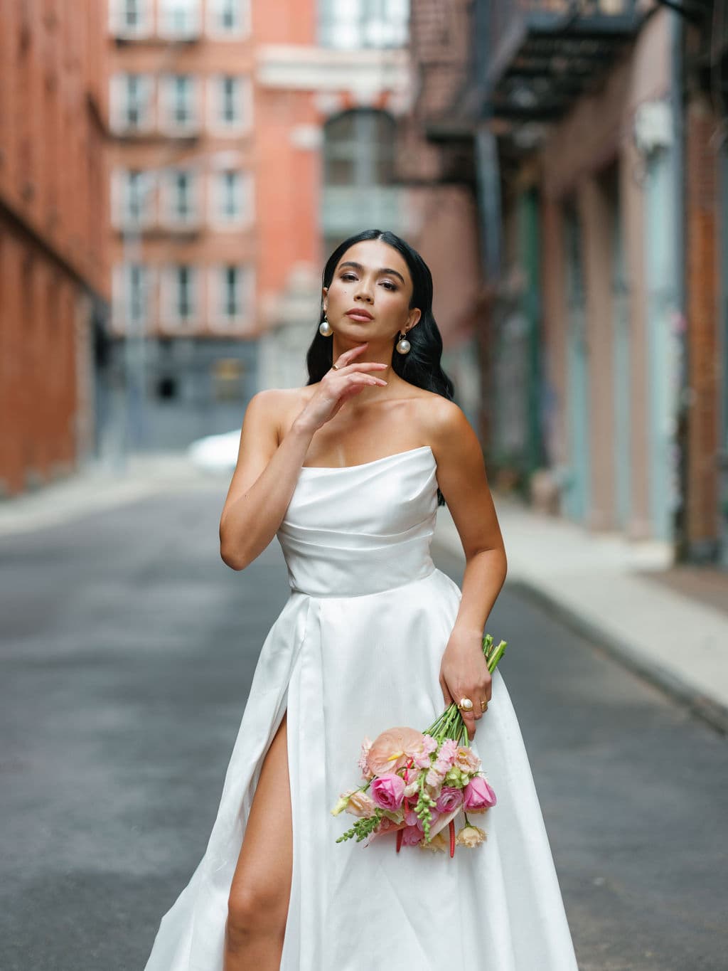 Shirin Hojayeva stands on a city street in a white strapless gown holding a pink bouquet, hand near her chin.